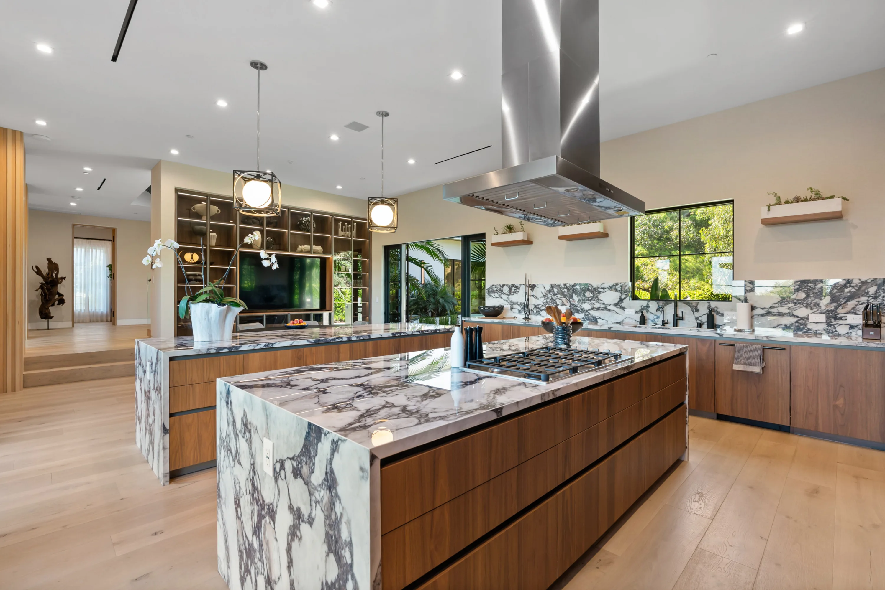 Twin kitchen islands in walnut and marble, gas cooktop under a stainless hood with garden windows behind