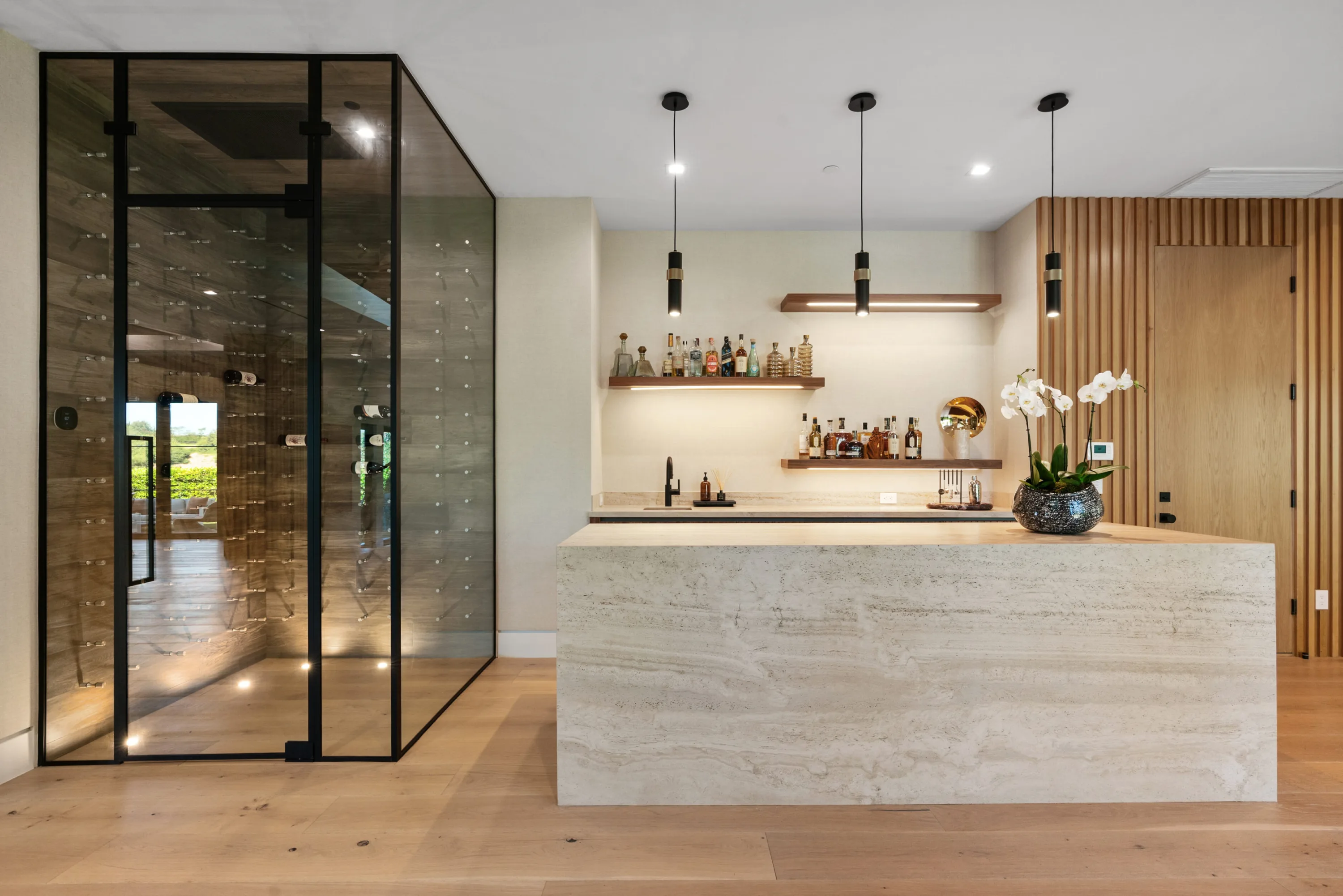 Travertine wet bar with backlit shelving next to a glass-walled wine room