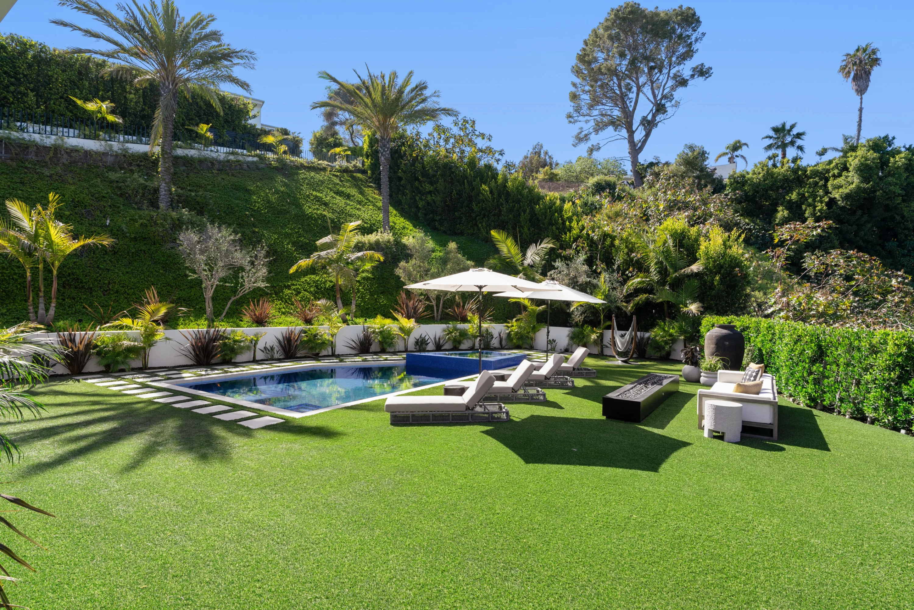 Pool and spa set into a broad lawn, sun loungers, fire table and a hammock under the hillside planting