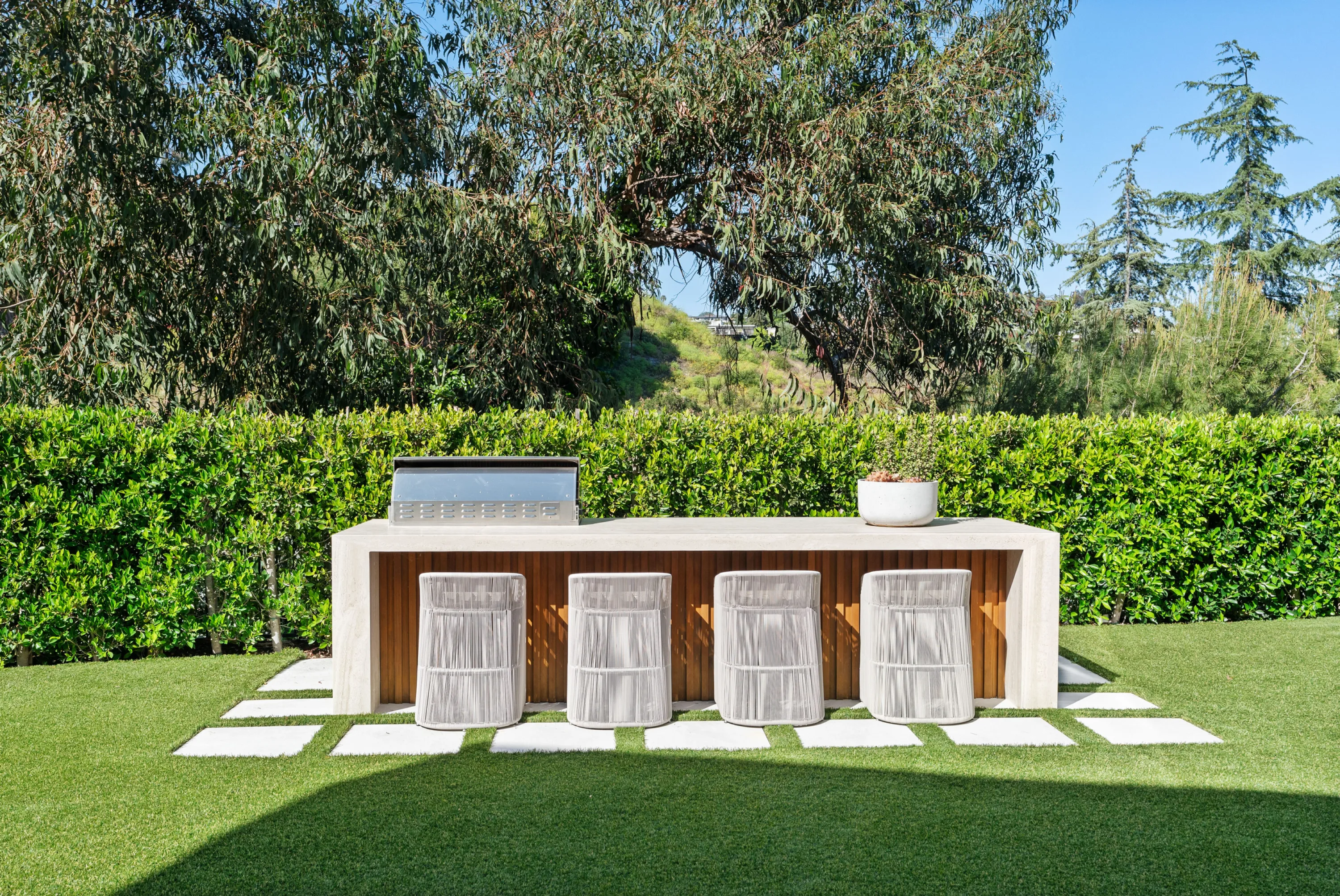 Outdoor bar counter with grill and four woven stools, backed by a clipped hedge and canyon trees