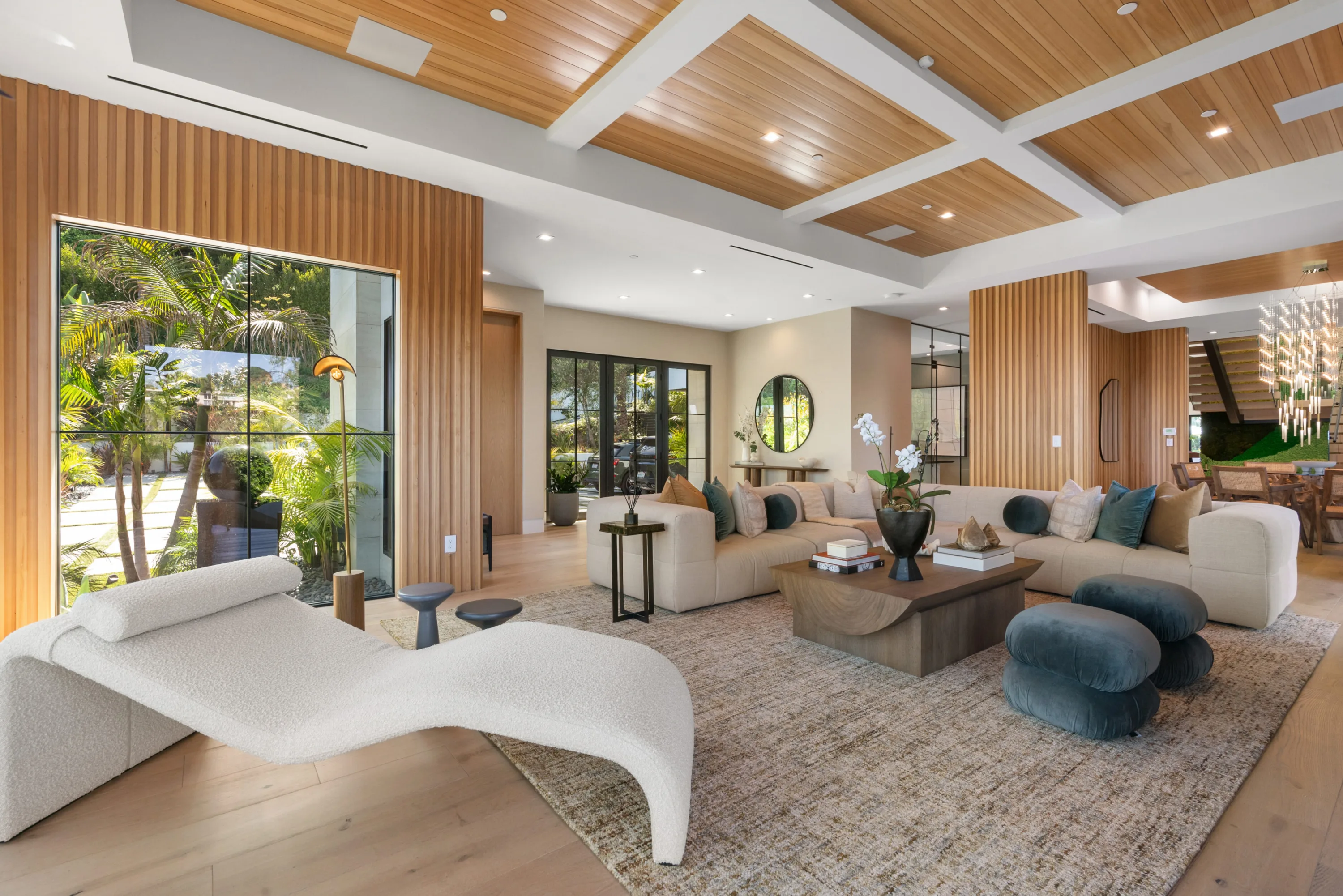 Great room under a cedar coffered ceiling, curved bouclé chaise by a tall window onto the palm garden