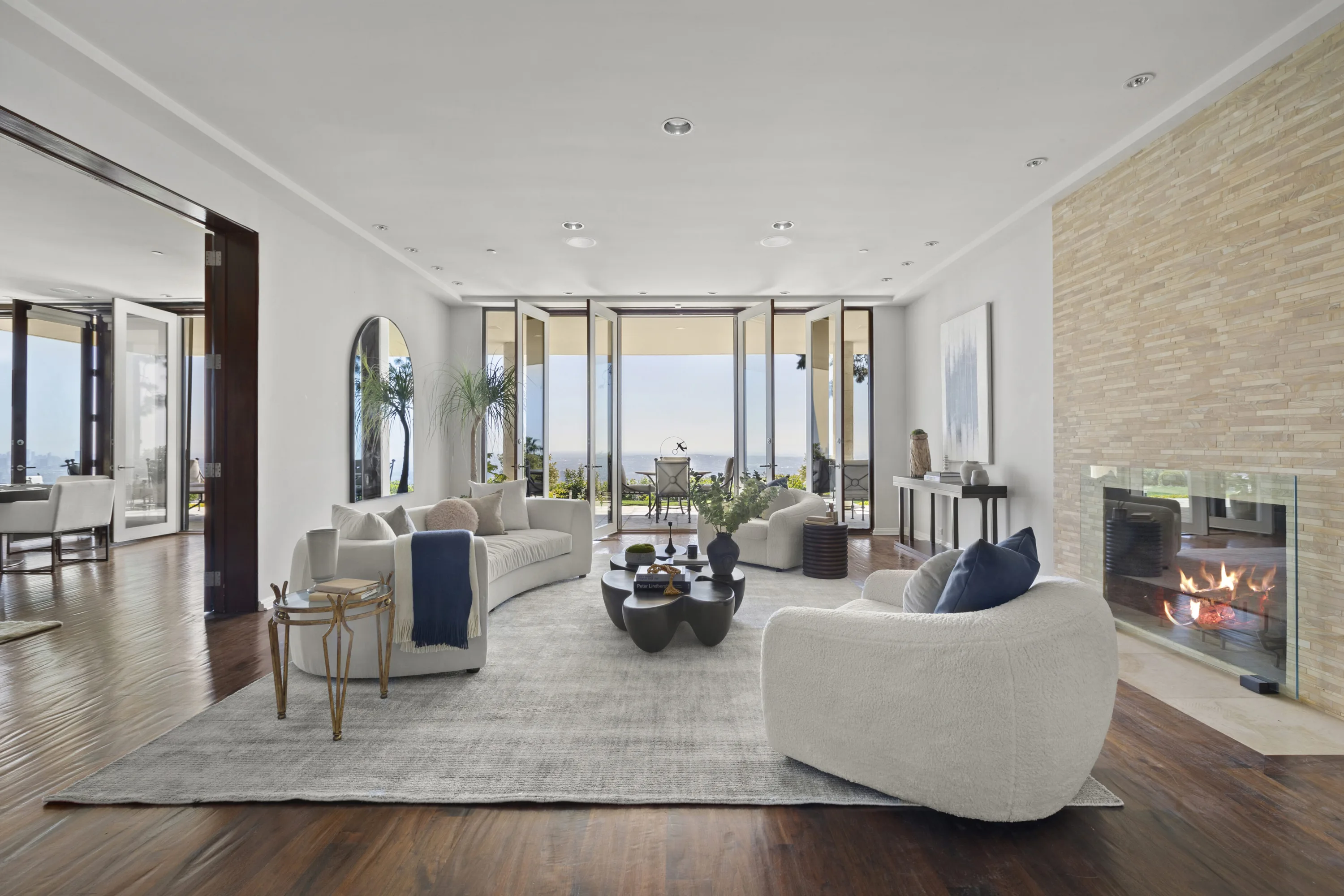 Living room with curved cream sofas, lit stone fireplace and folding glass walls opening to a hazy city horizon
