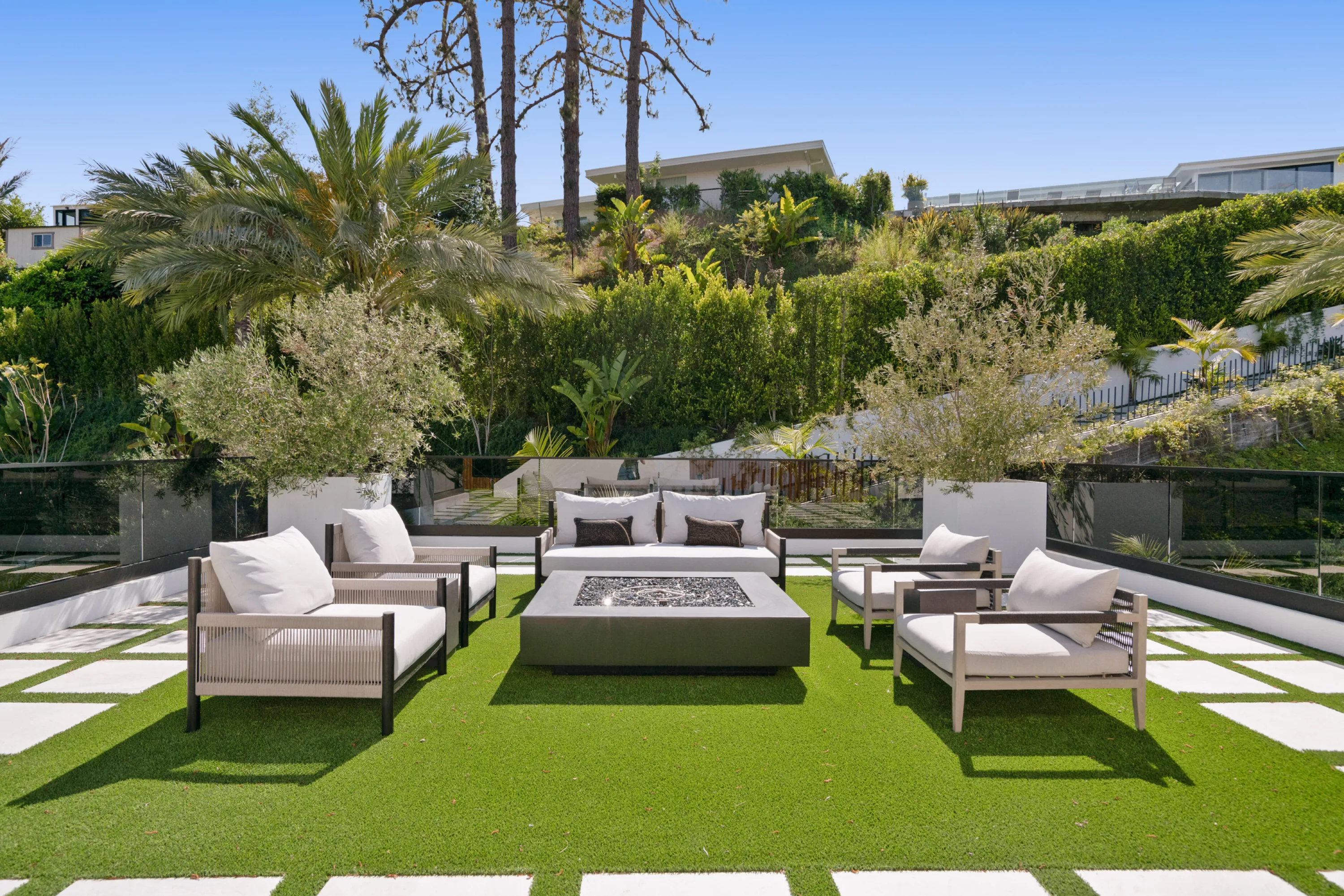 Lawn terrace fire pit ringed by cushioned sofas and armchairs, hedged hillside and palms beyond