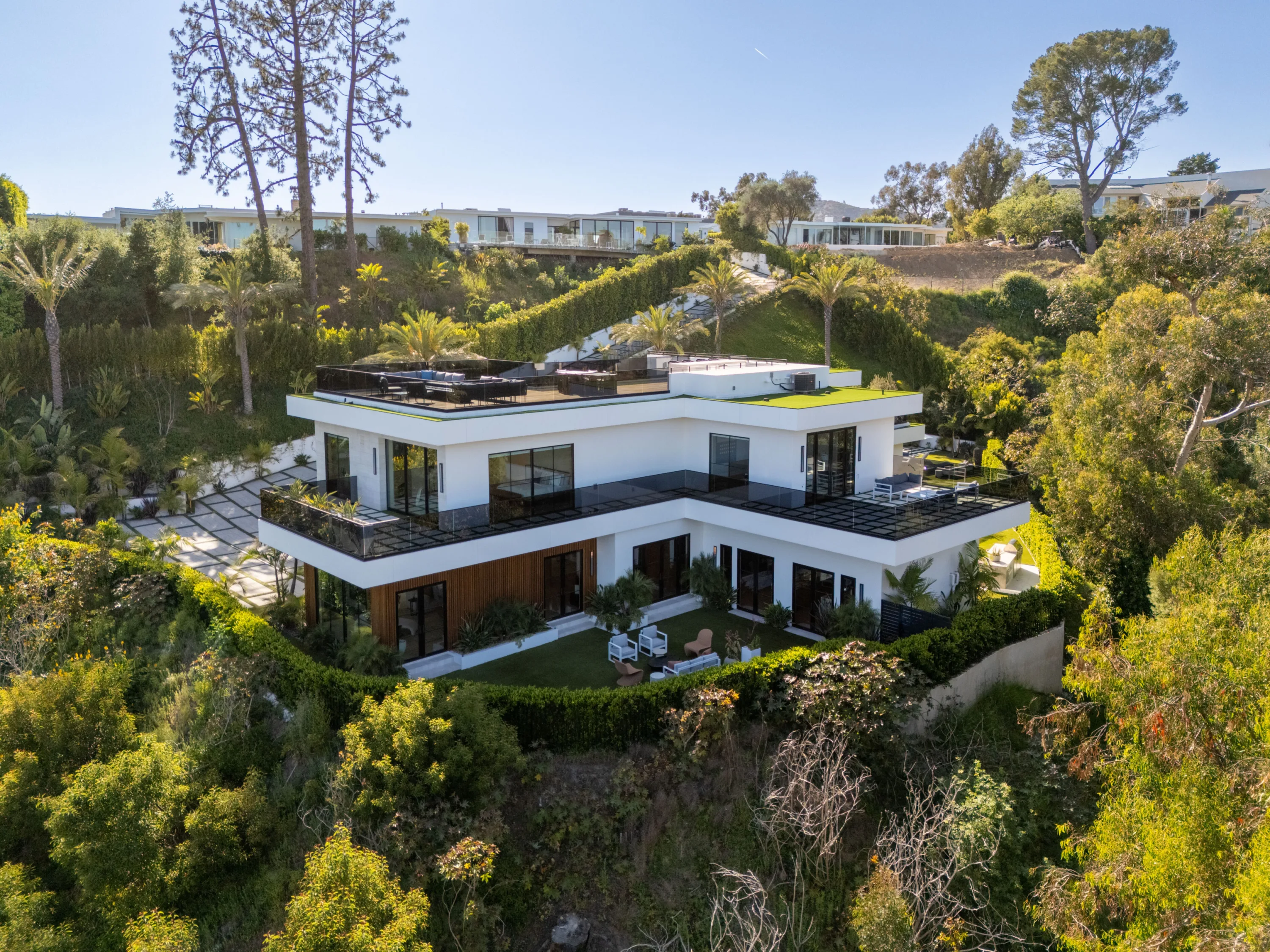Aerial rear view of the white stacked-volume house, glass-railed terraces and lawn tucked into the canyon