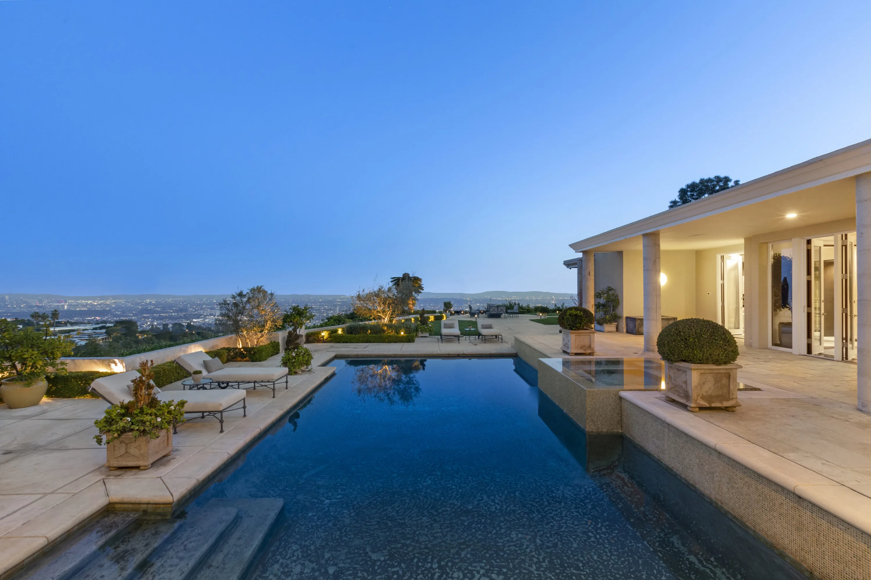 Resort pool and stone sun loungers at dusk, city lights stretching to the horizon beyond the terrace