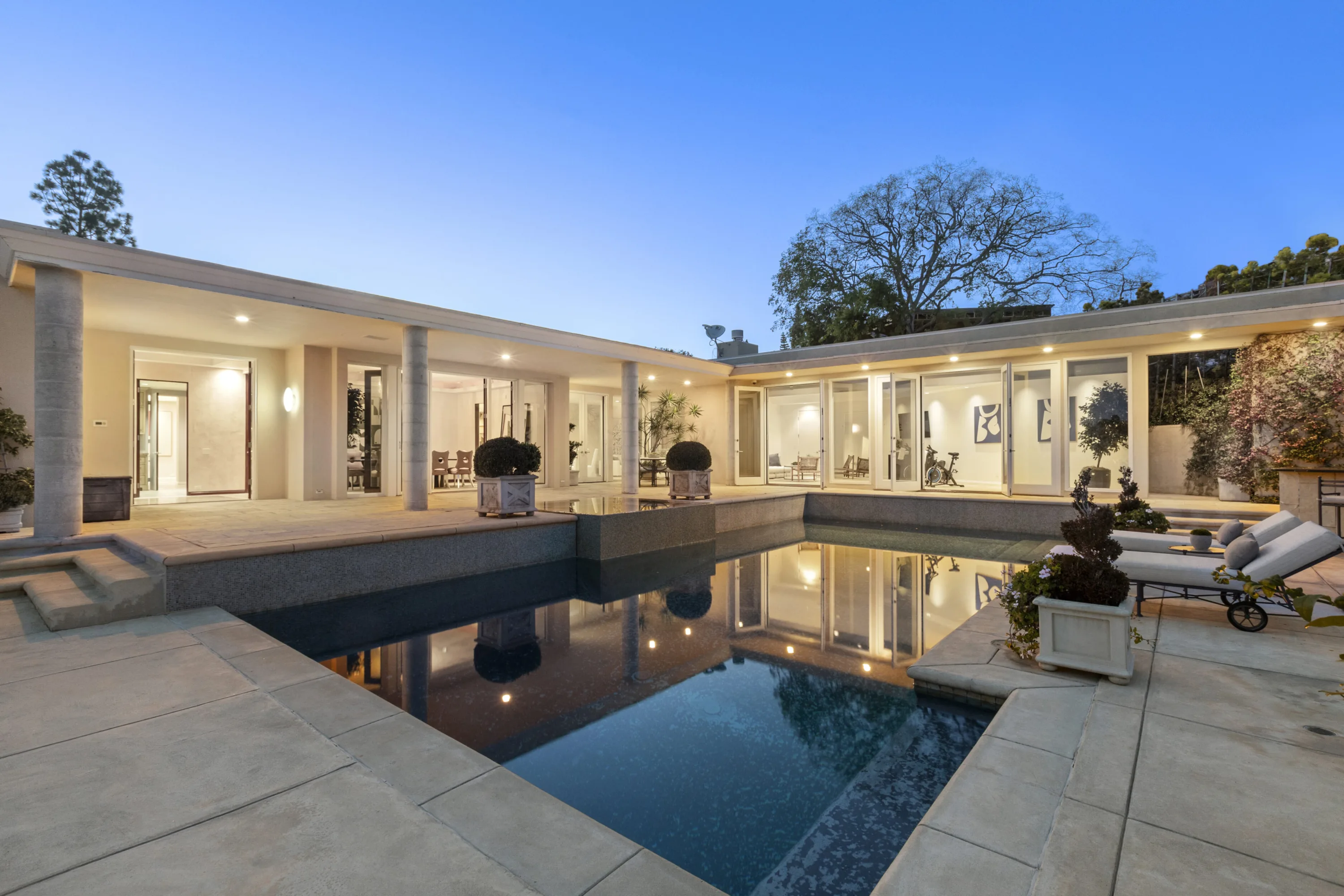 Pool wrapped by the single-storey glass wings, lit rooms and colonnade reflected in still water at blue hour