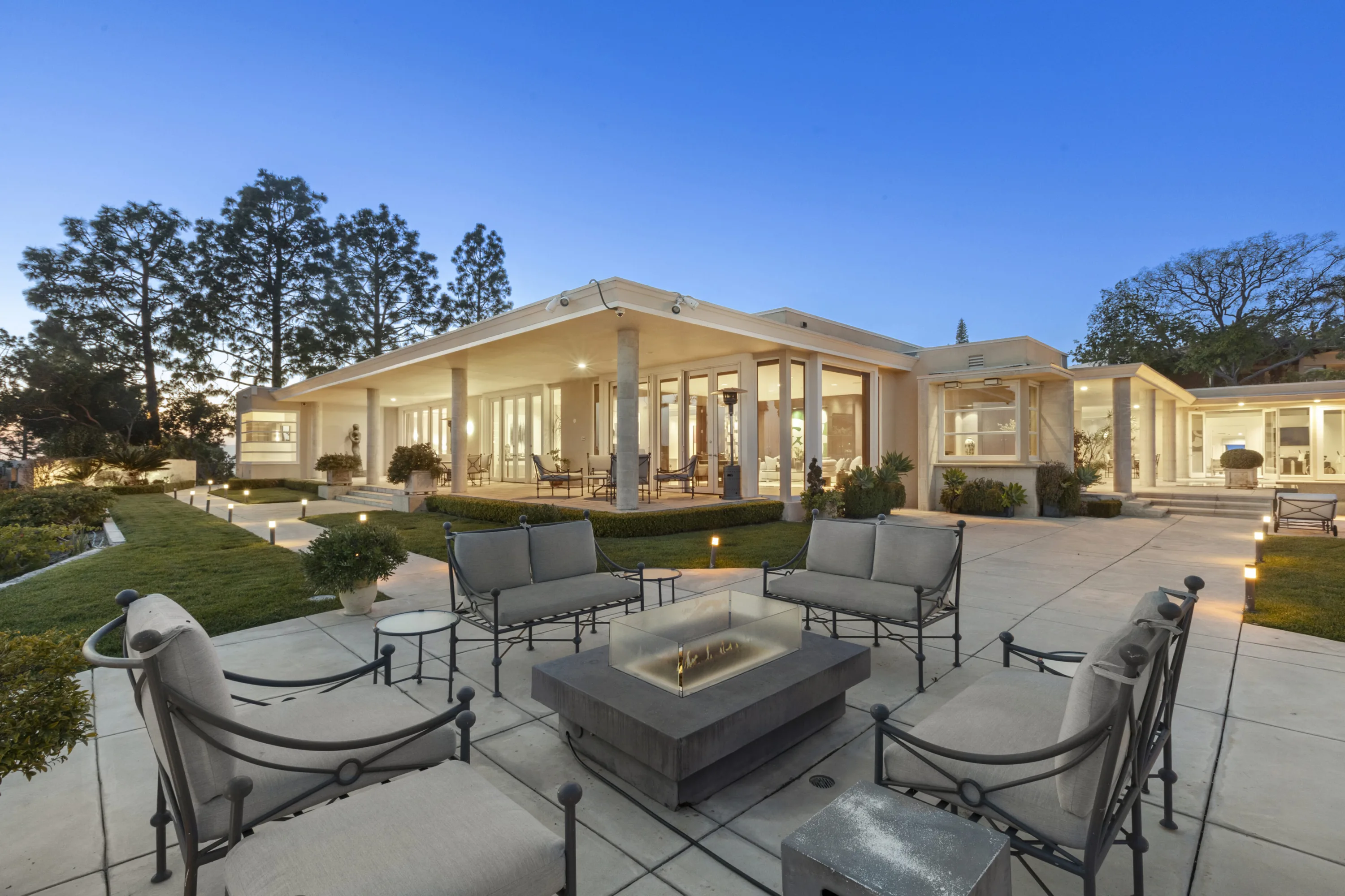 Fire table ringed by cushioned patio armchairs on the lawn terrace, covered loggia glowing behind