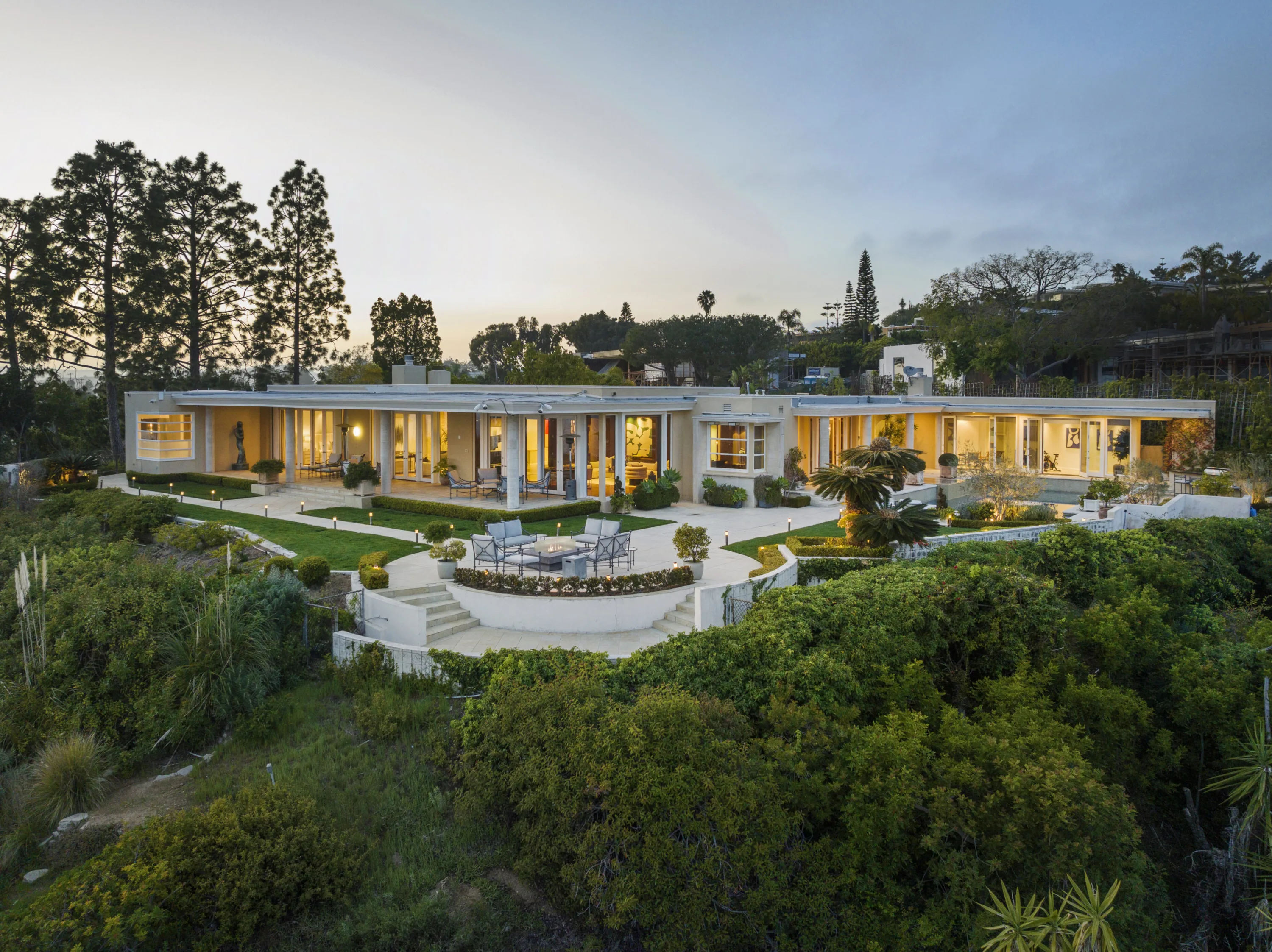 Elevated view of the long glass facade, curved fire-pit terrace and lawn above a brushy canyon slope