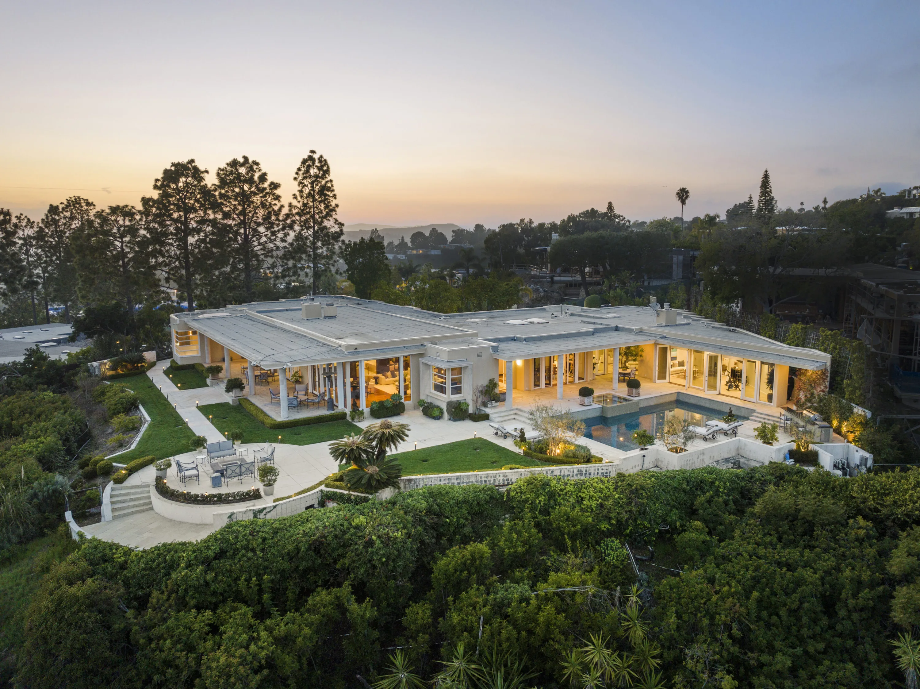 Drone view at golden hour showing the full wingspan of the house, lawns, pool deck and lit interiors
