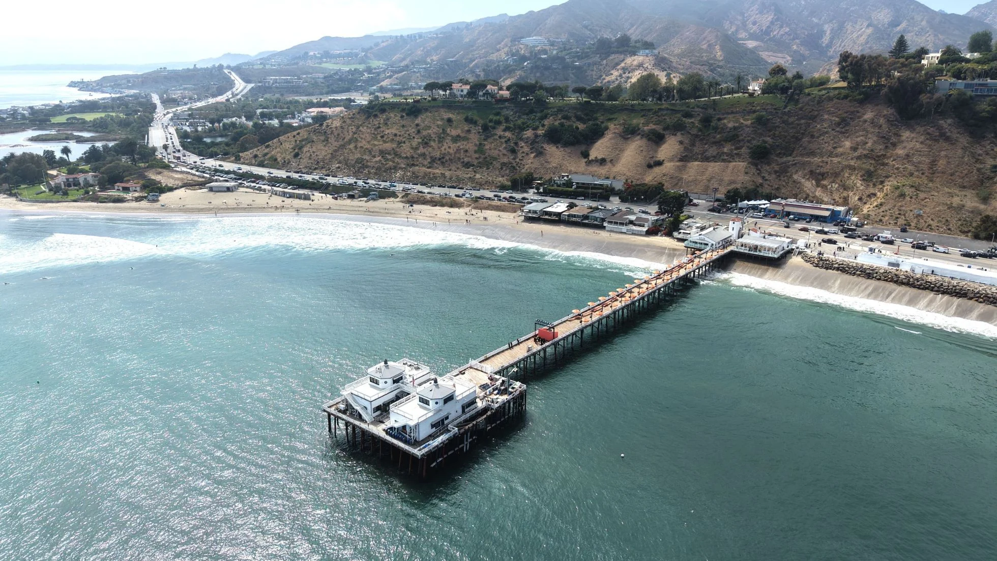 Aerial view of Malibu Pier extending into the Pacific Ocean with the coastline and Santa Monica Mountains behind it.