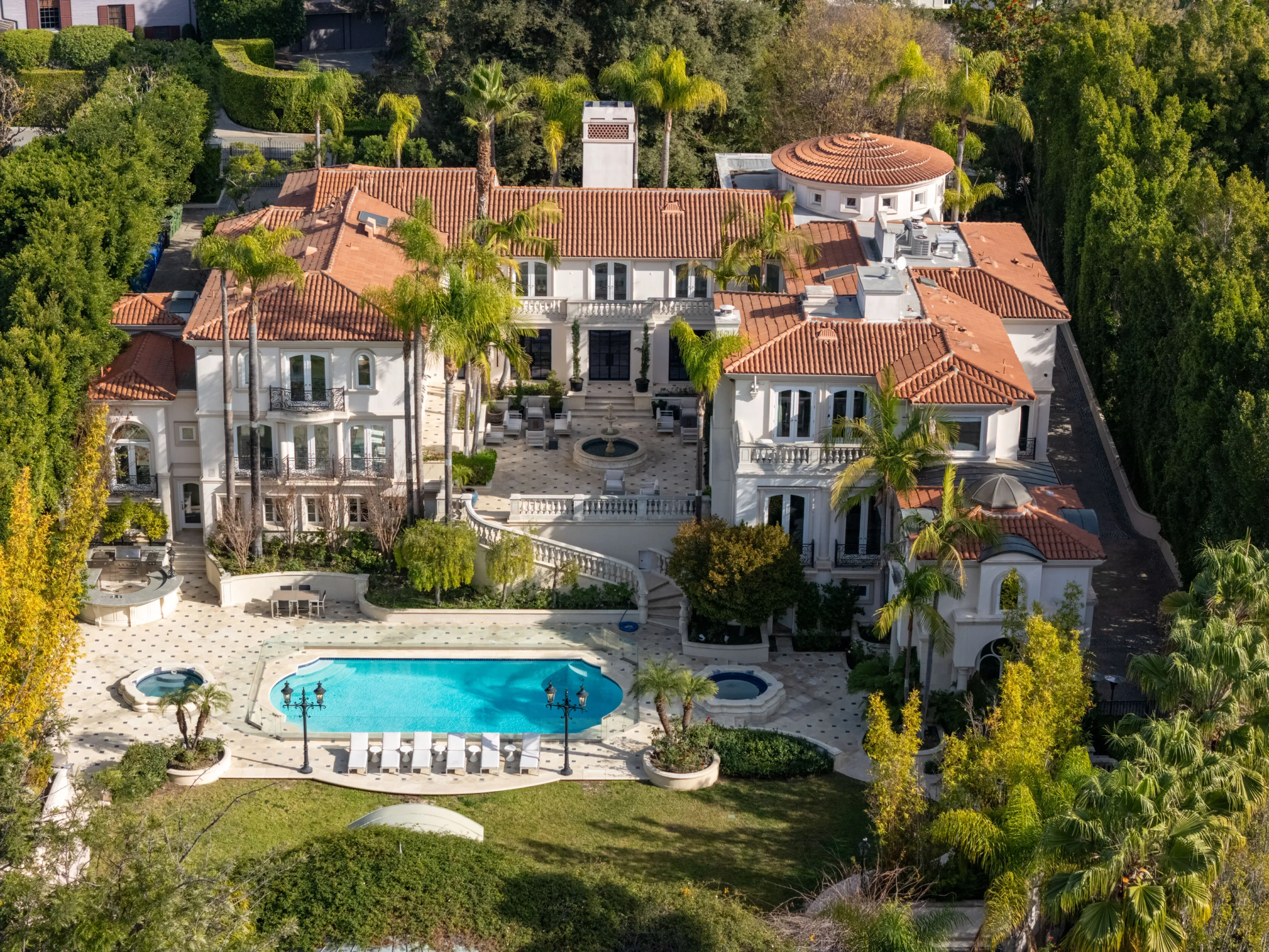 Aerial view of a tile-roofed Mediterranean estate with tiered terraces, fountain courtyard and oval pool flanked by two spas