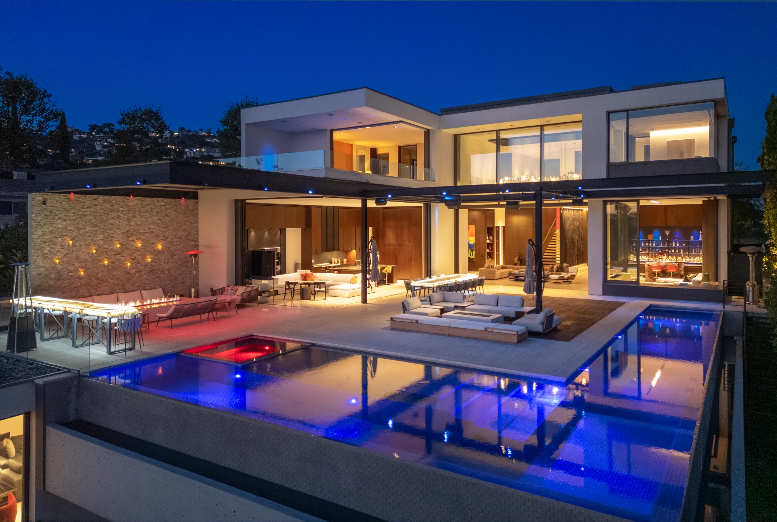 Blue-lit pool wrapping the hillside facade at blue hour, walls of glass folded open to the deck