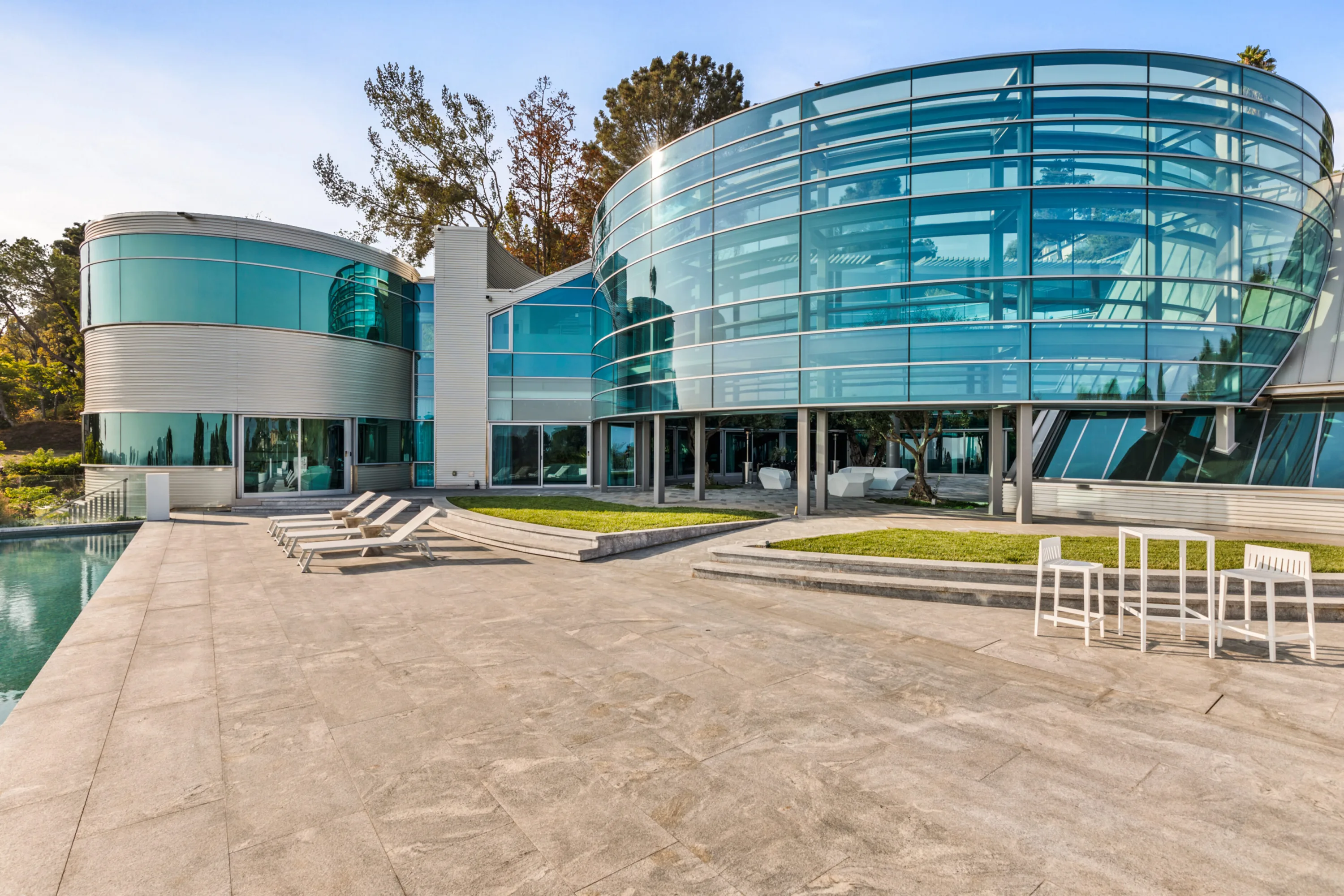 Curved glass-and-steel facade of Jetson Estate seen from the stone pool terrace, loungers lined along the water