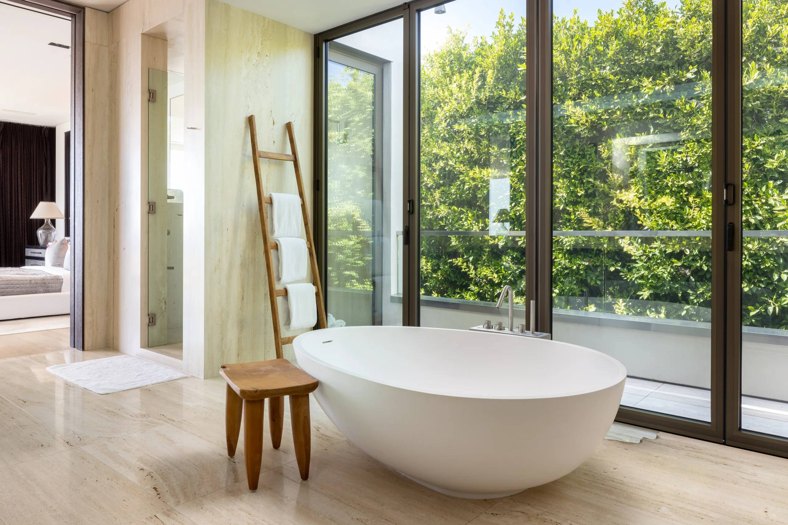 Sculptural freestanding tub in a travertine ensuite, framed by glass and a wall of greenery