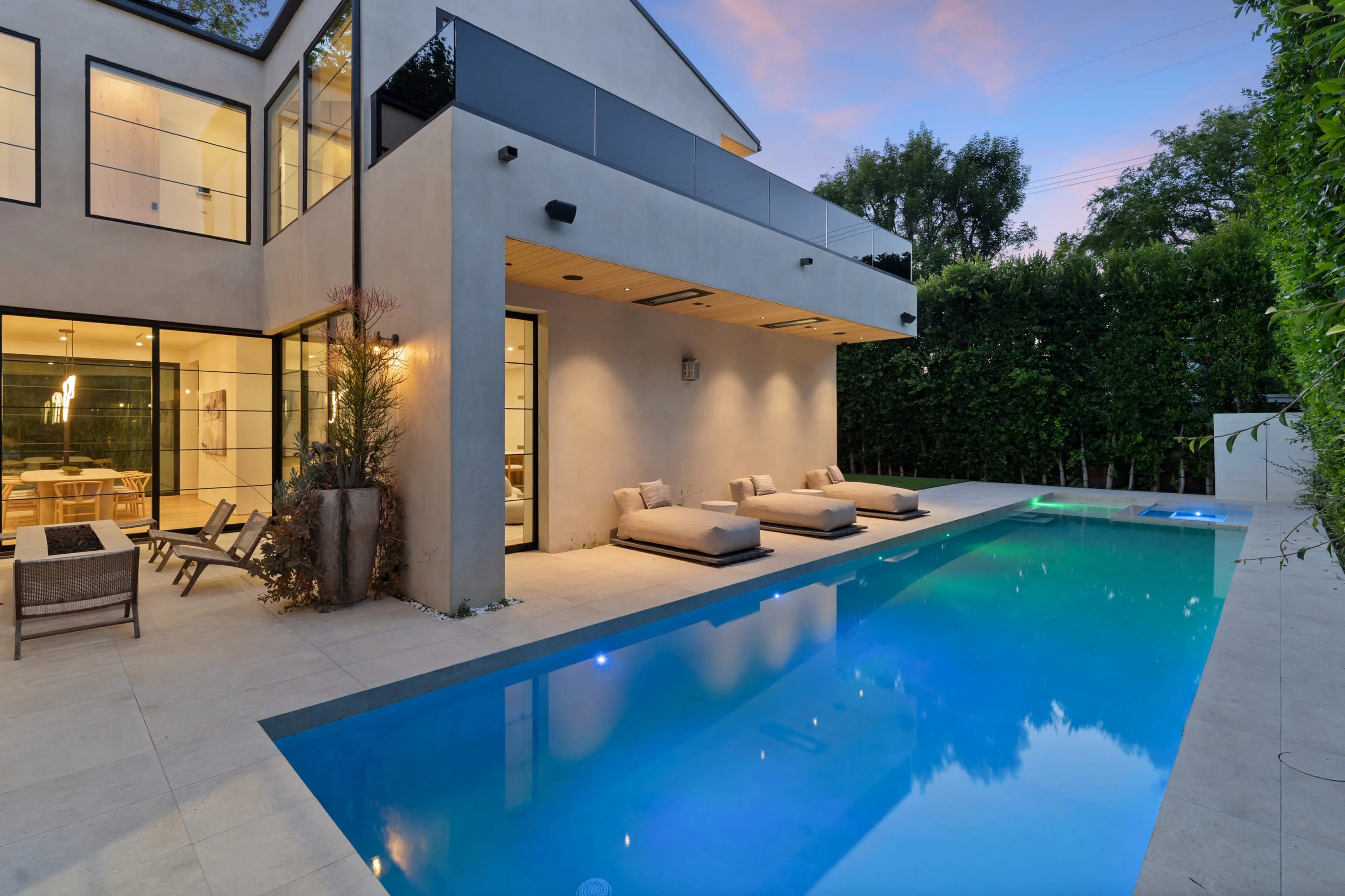 Lit lap pool at dusk with twin daybeds under a cantilevered terrace and glowing dining room beyond