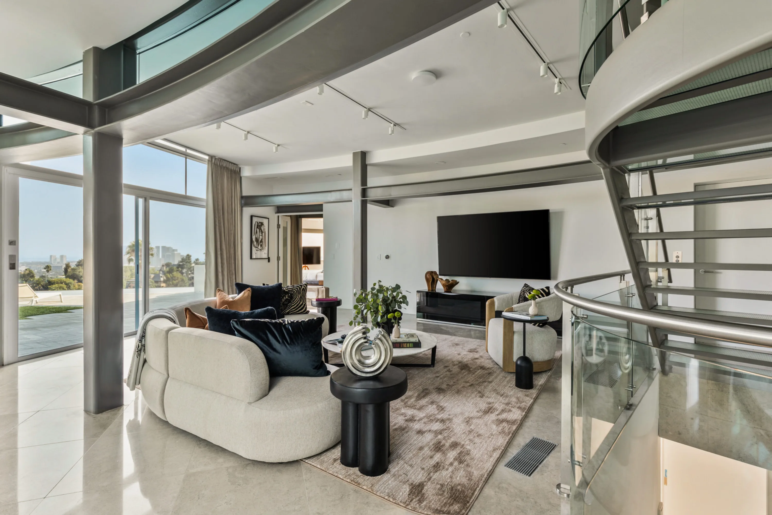 Upper lounge with curved sofa, wall-mounted TV and a glass stair rail, terrace and skyline through the windows