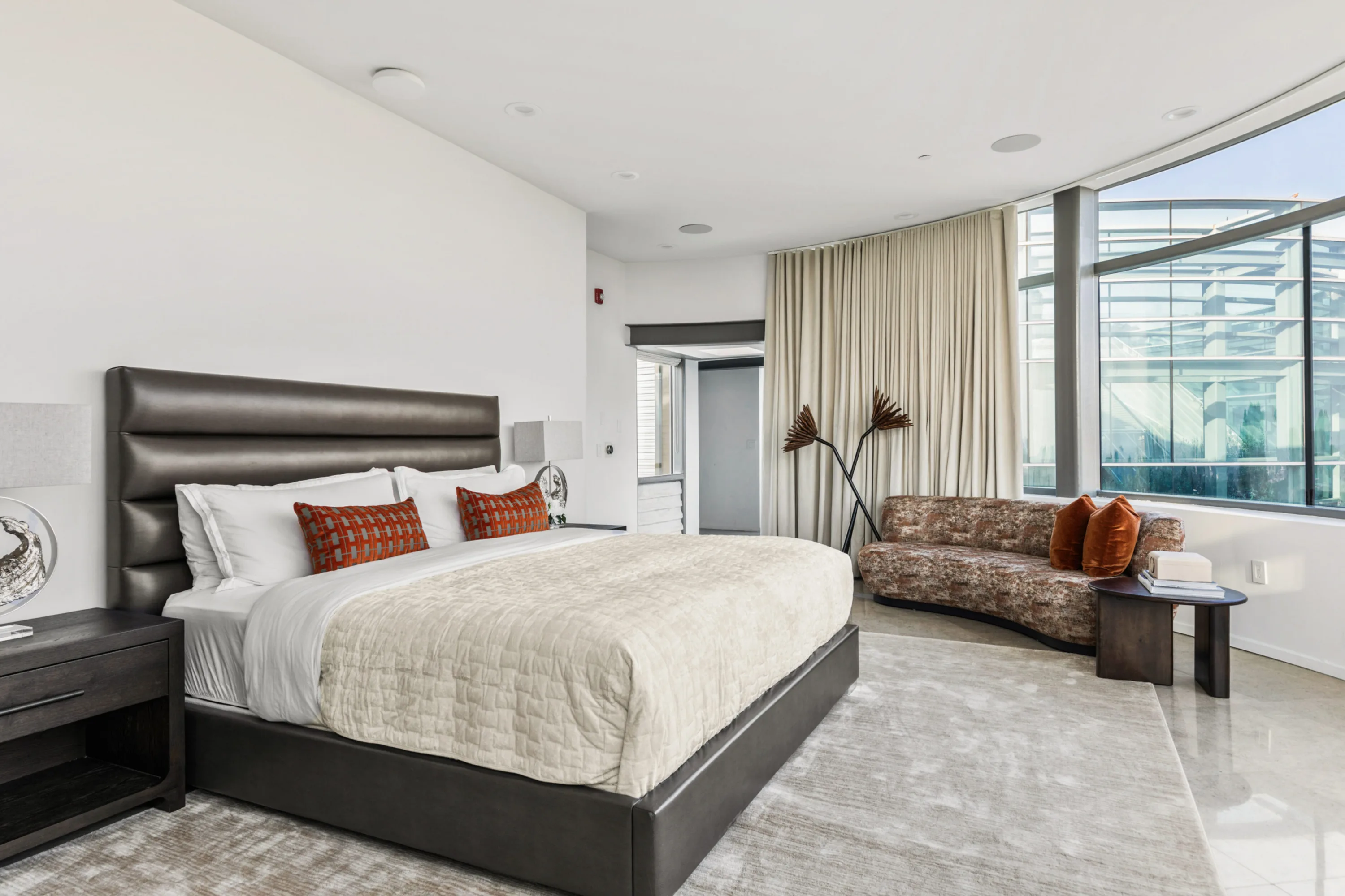 Guest bedroom with leather-panelled king bed, rust cushions and a curved window wall framing the glass wing