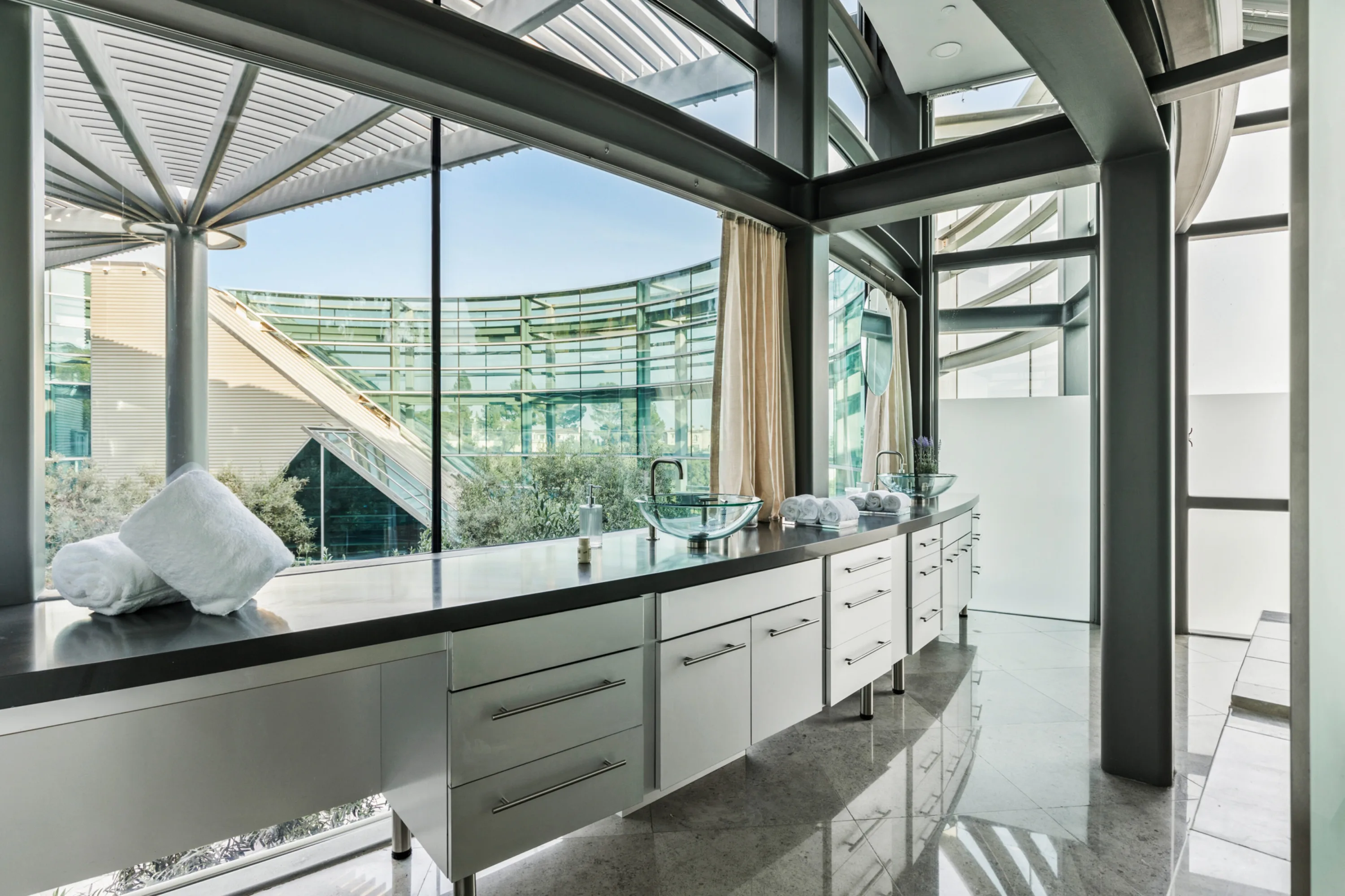Double-vanity bathroom with glass vessel basins and rolled towels, full-height windows onto the curved facade