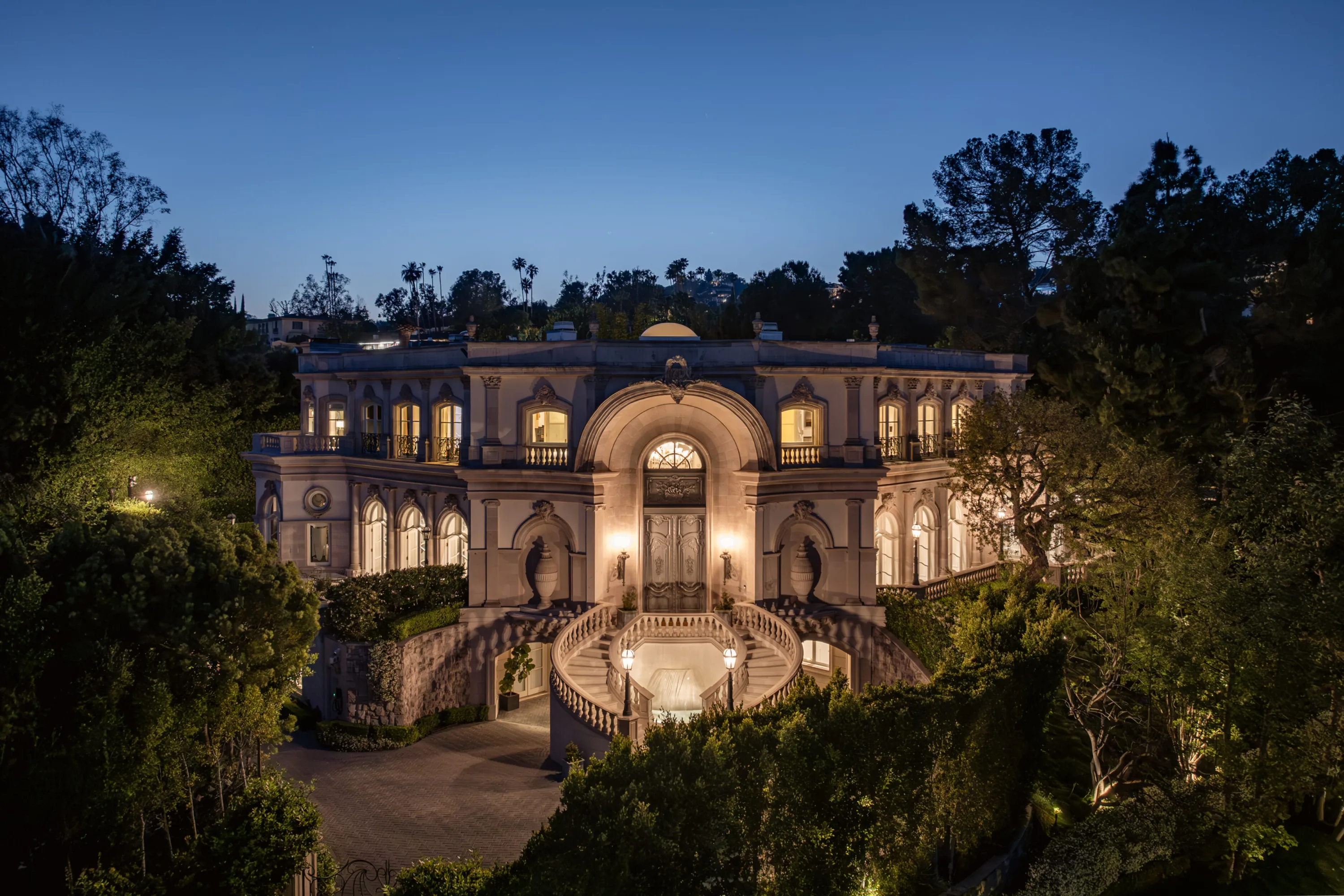 Neoclassical villa facade lit at dusk, its arched entry and twin curving staircases glowing above a fountain