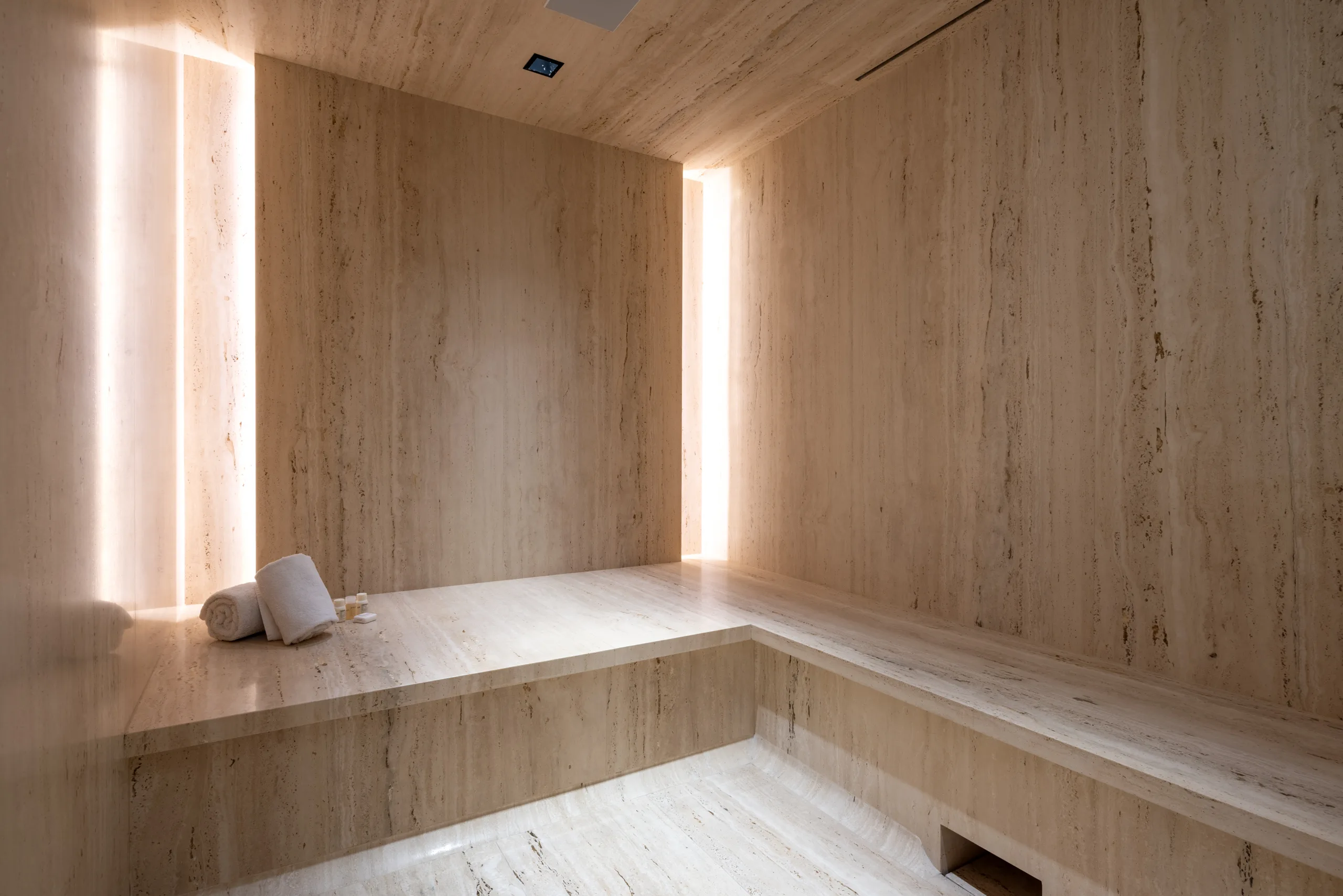 Travertine steam room with benches, rolled towels and slim backlit light slots