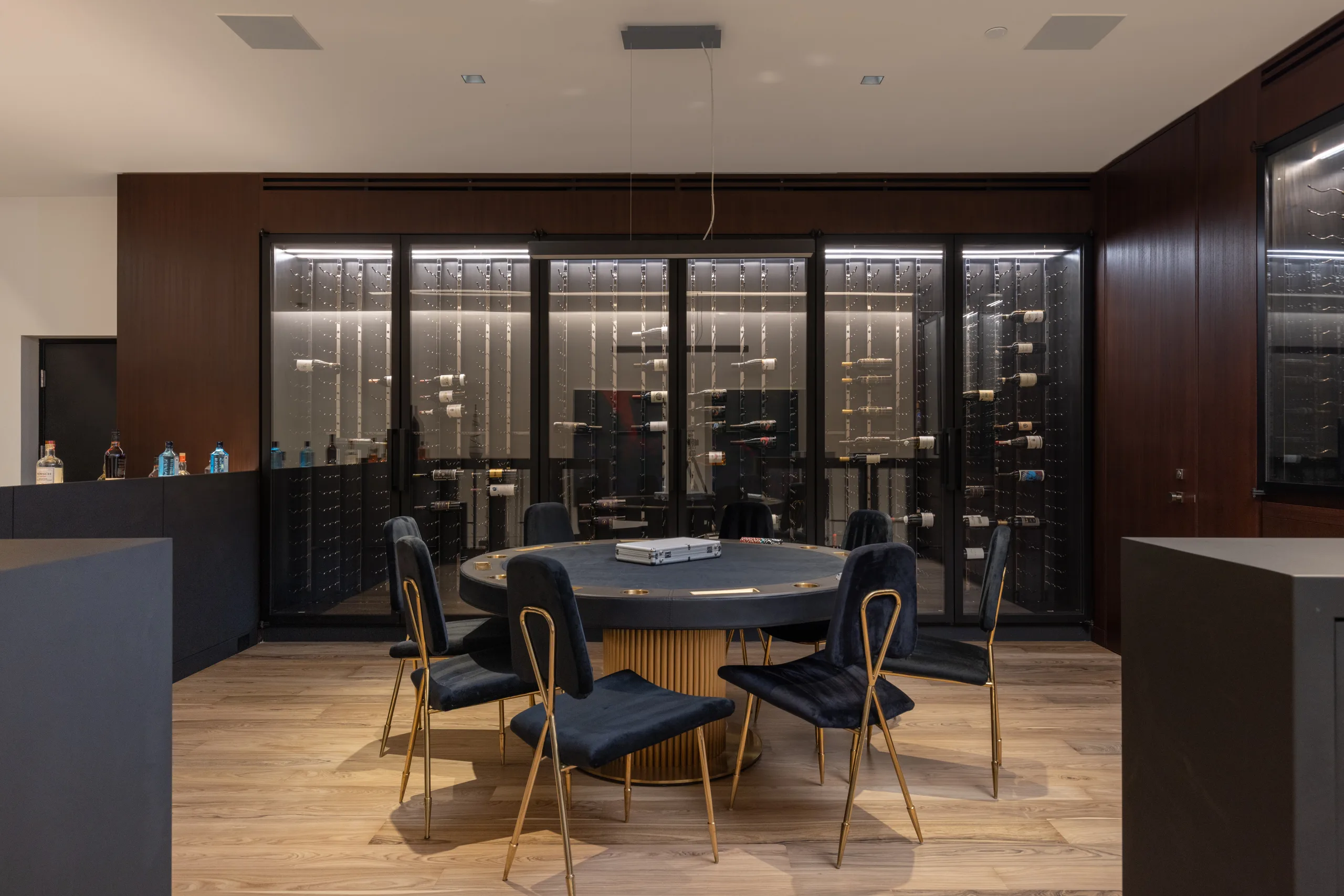 Poker table ringed by velvet chairs in front of a glass-walled wine cellar and bar counter