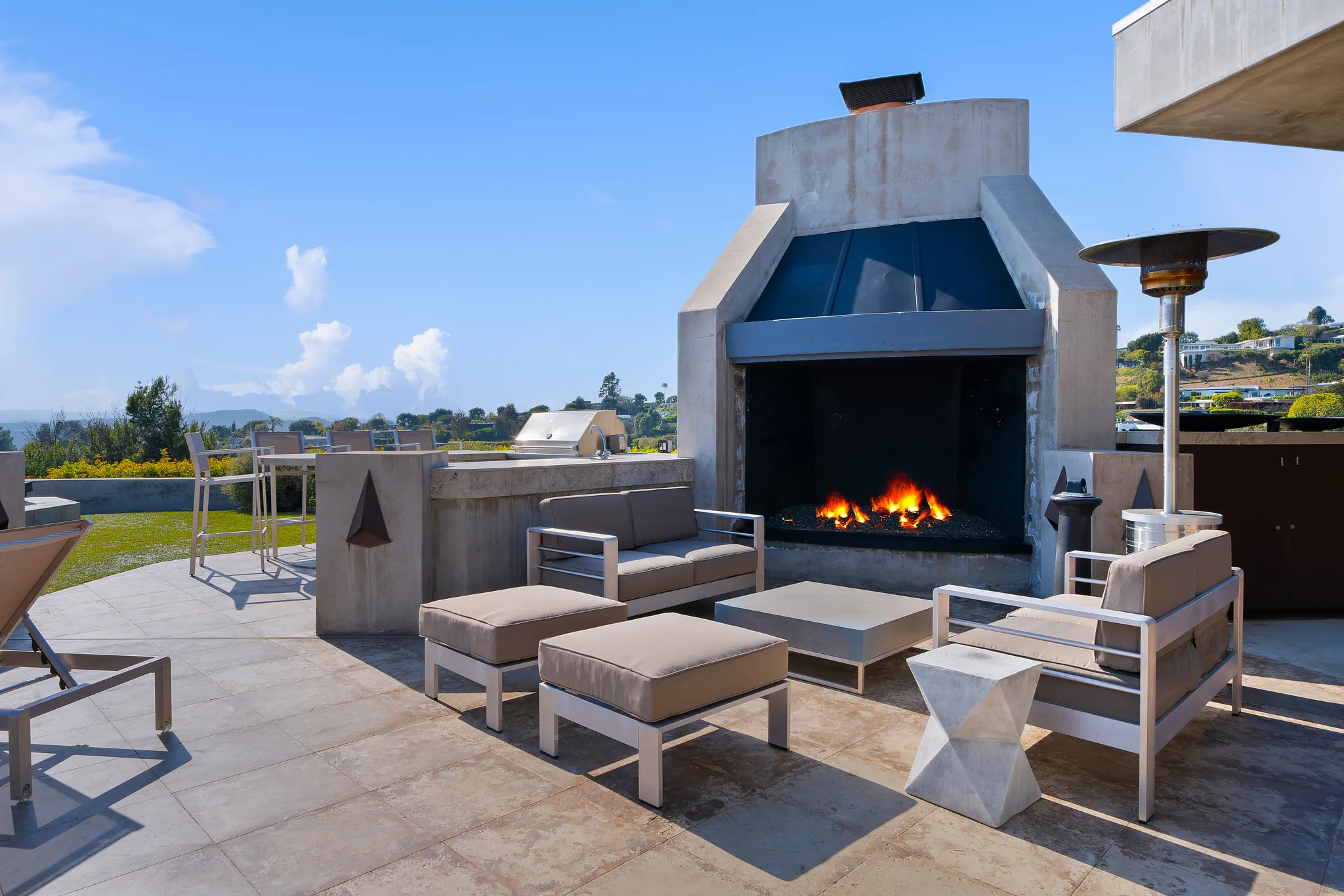 Outdoor lounge and built-in grill beside a tall concrete fireplace with flames, patio heater alongside