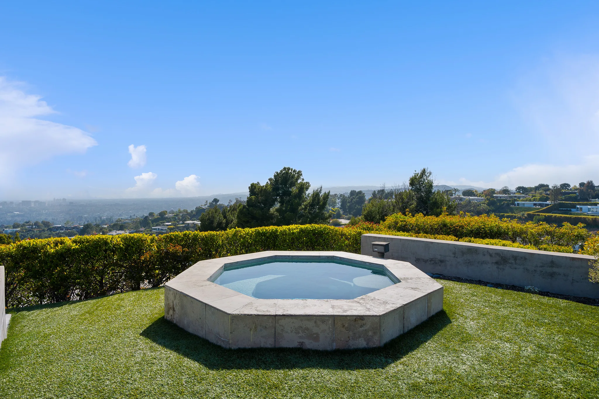 Octagonal stone-rimmed spa set into lawn, hedges opening to a hazy city panorama