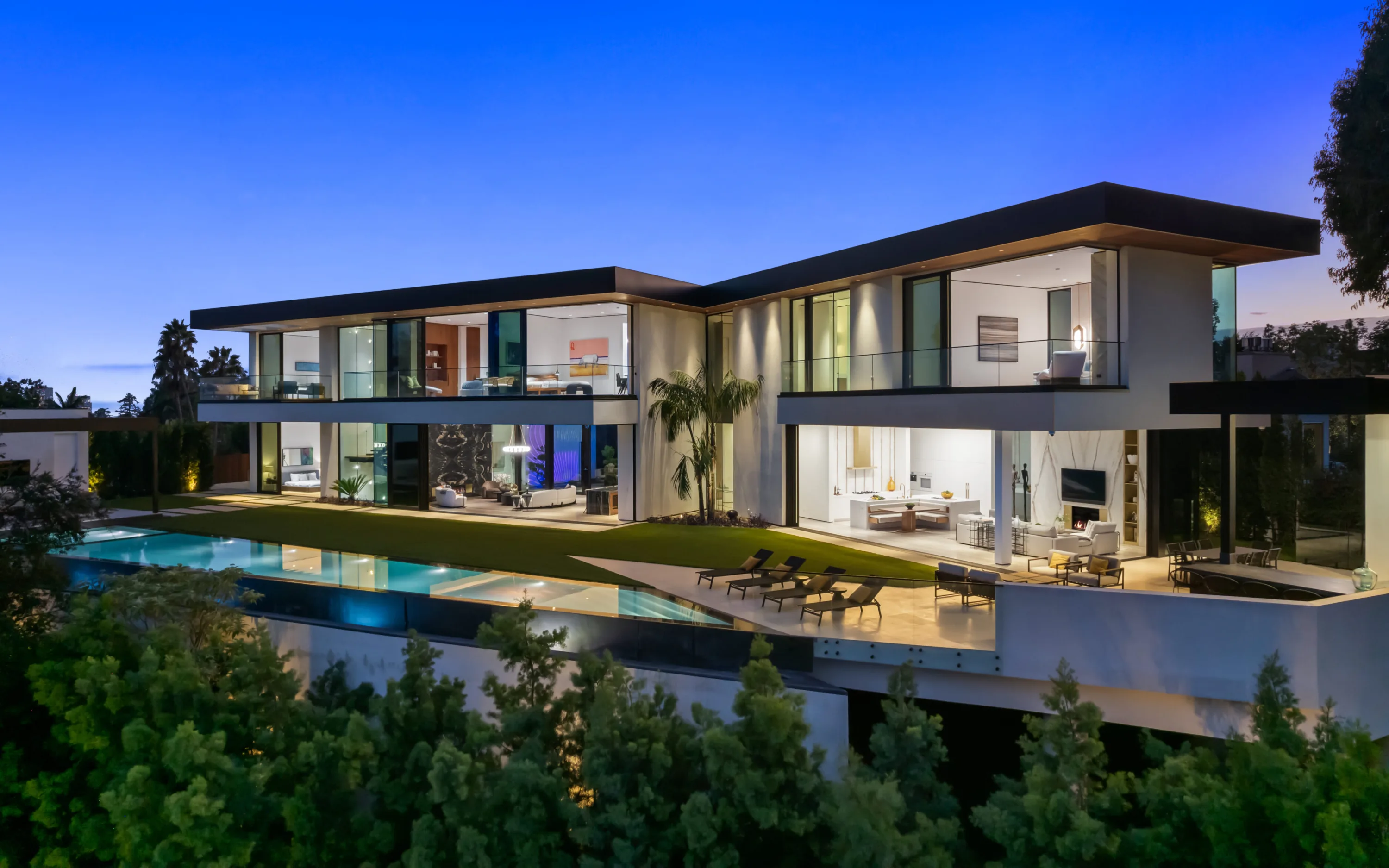 Glass-walled contemporary villa lit at dusk above a hillside, with lap pool, lawn terrace and open living rooms