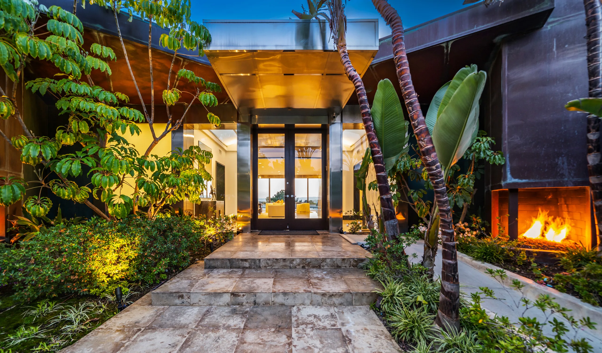 Entrance courtyard at twilight, travertine path to glass front doors with a lit fire feature to the right