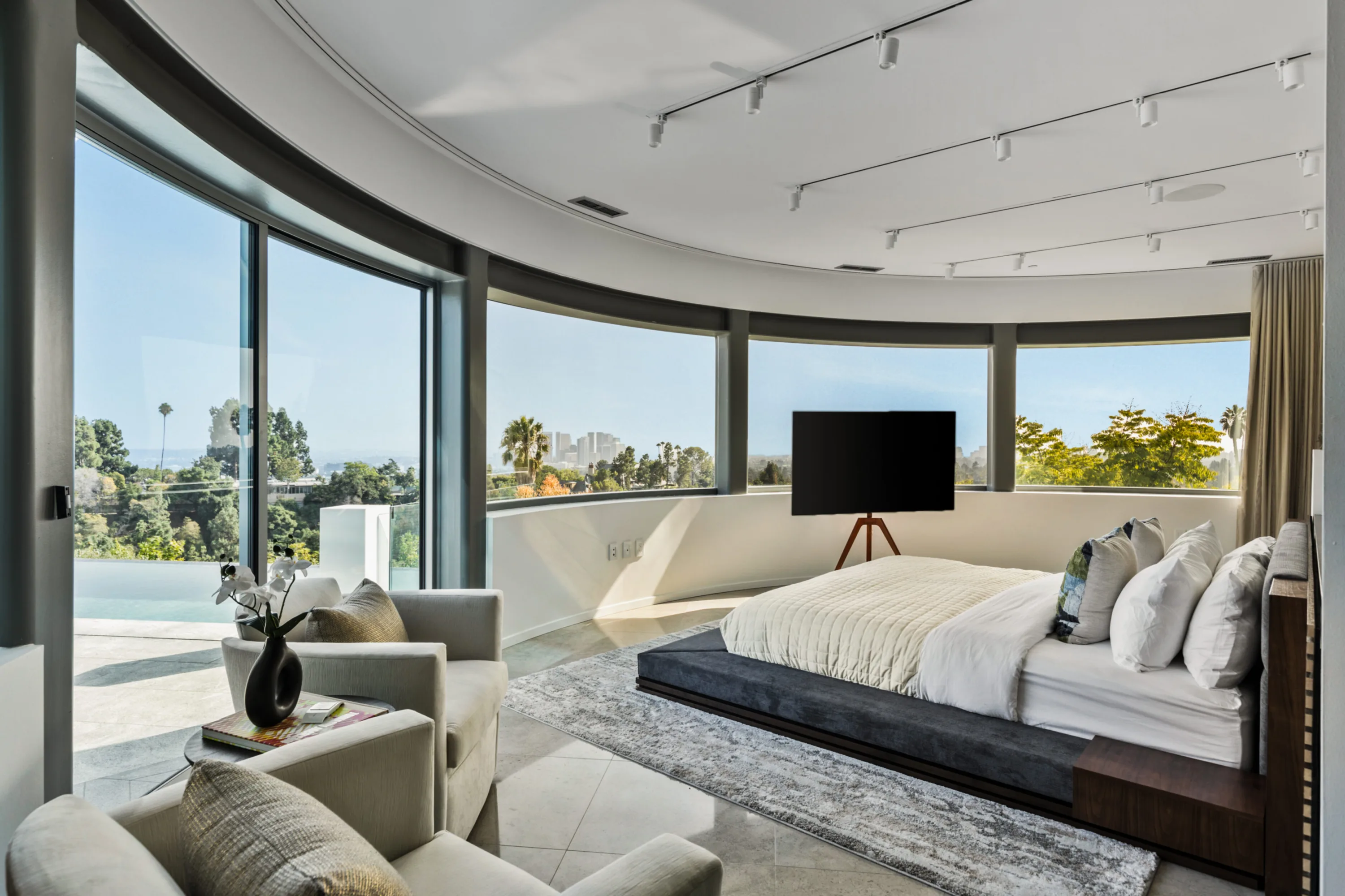 Primary suite curving around floor-to-ceiling glass, platform bed and armchairs facing the pool and city haze