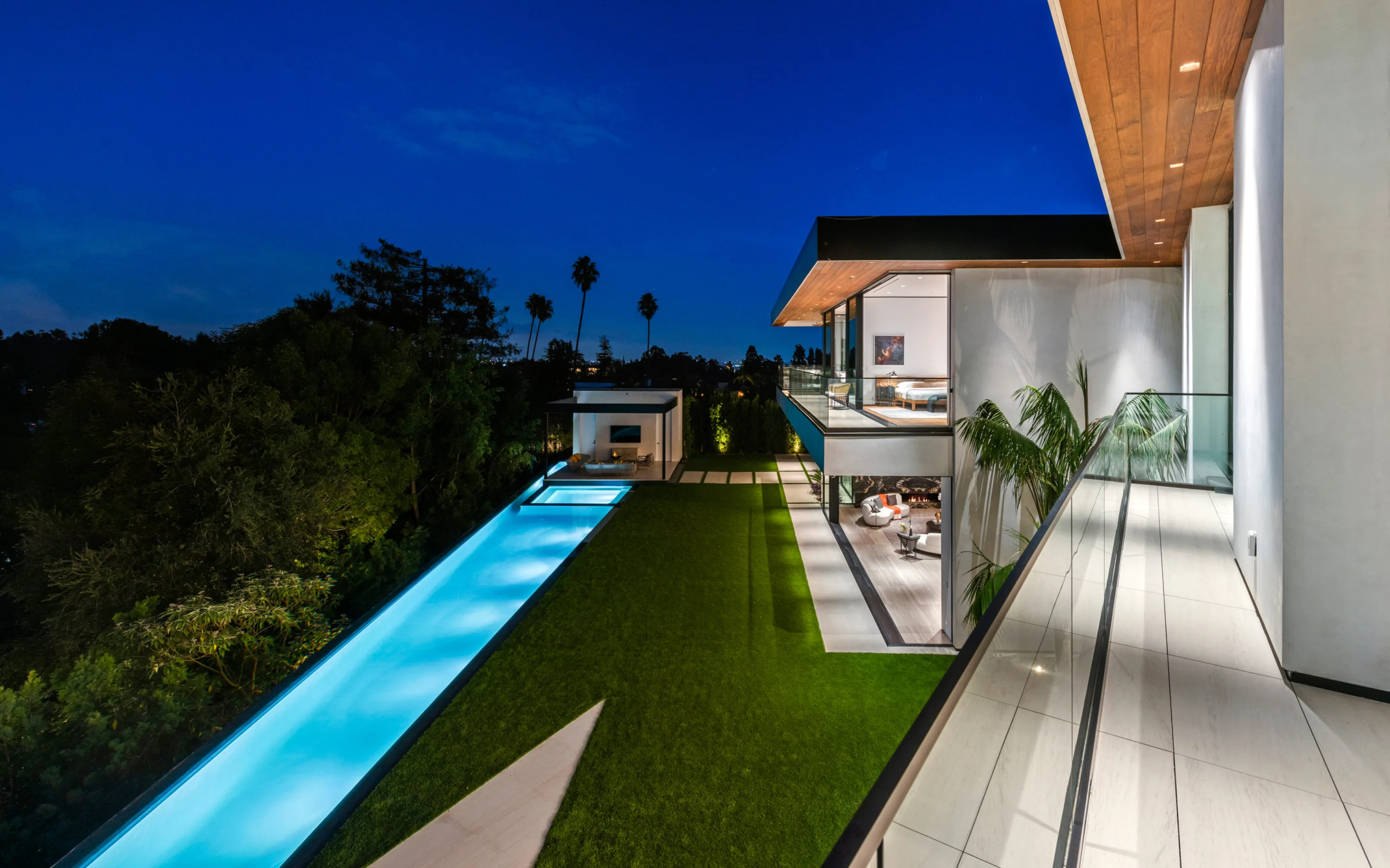 Lit lap pool and lawn seen from a glass-railed terrace at blue hour, with the villa's glowing living room beyond