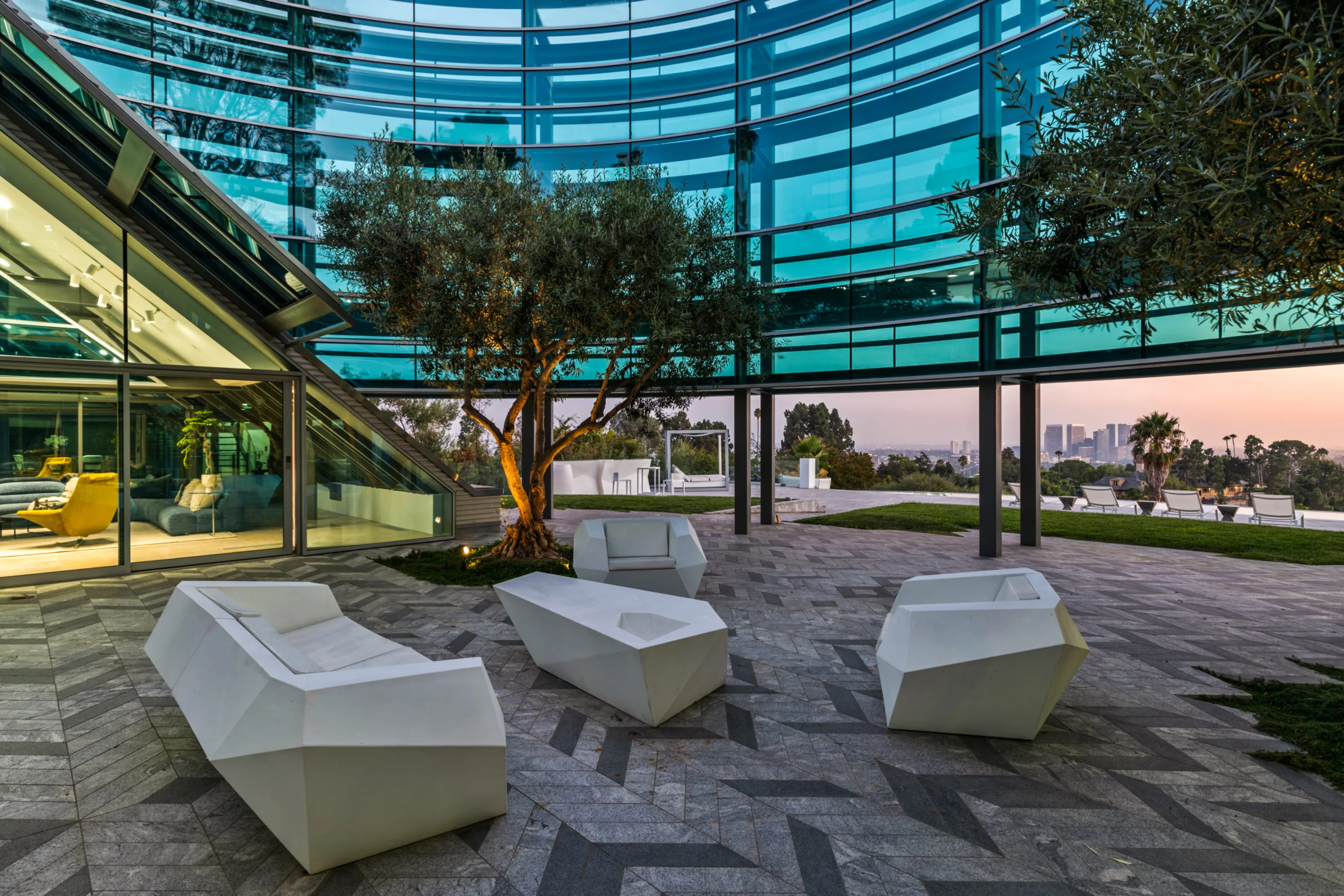 Courtyard at dusk with faceted white loungers around a lit olive tree, skyline glowing past the glass curve