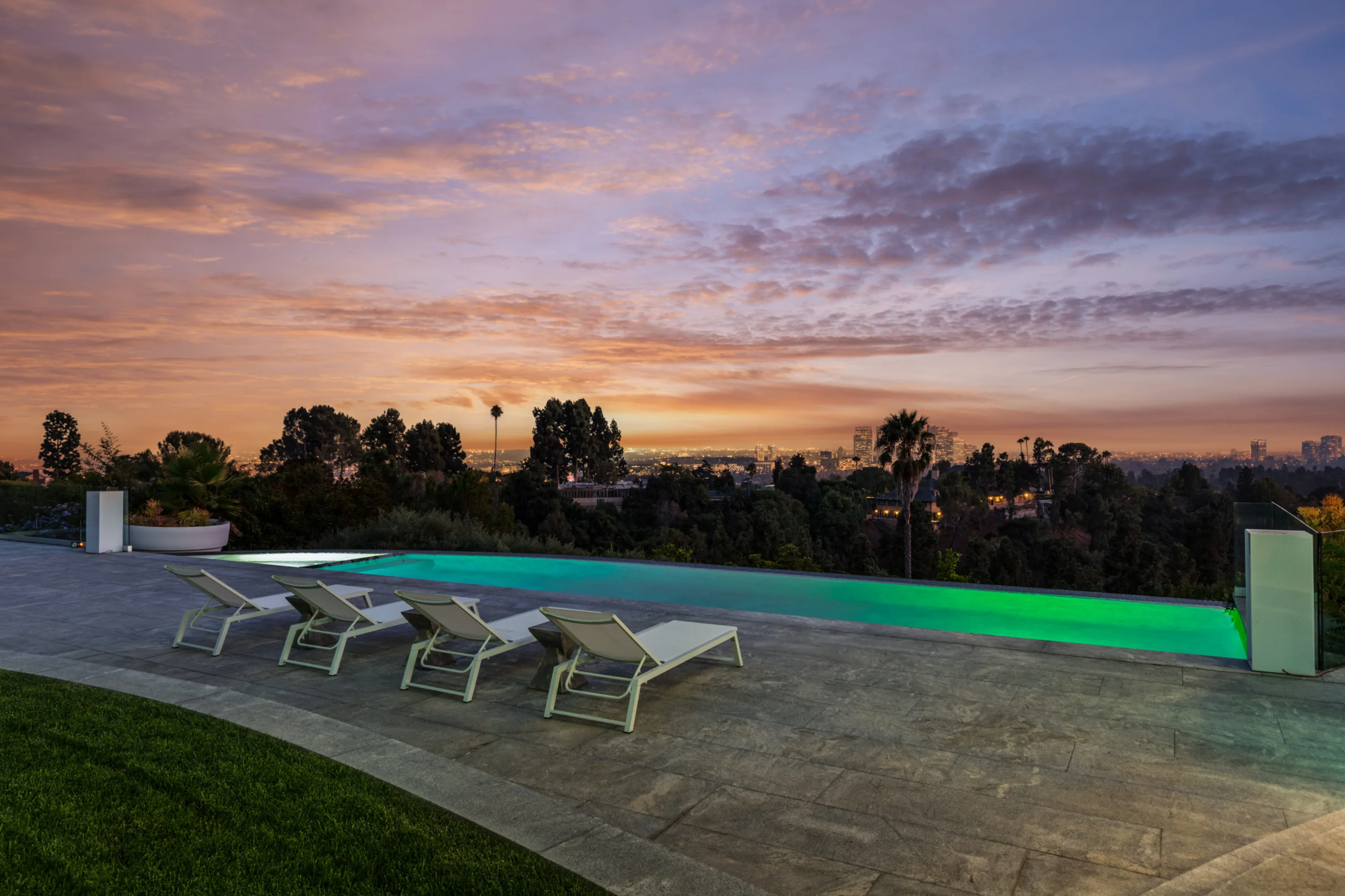 Lit infinity pool glowing green against a pink sunset, city lights spread below the terrace