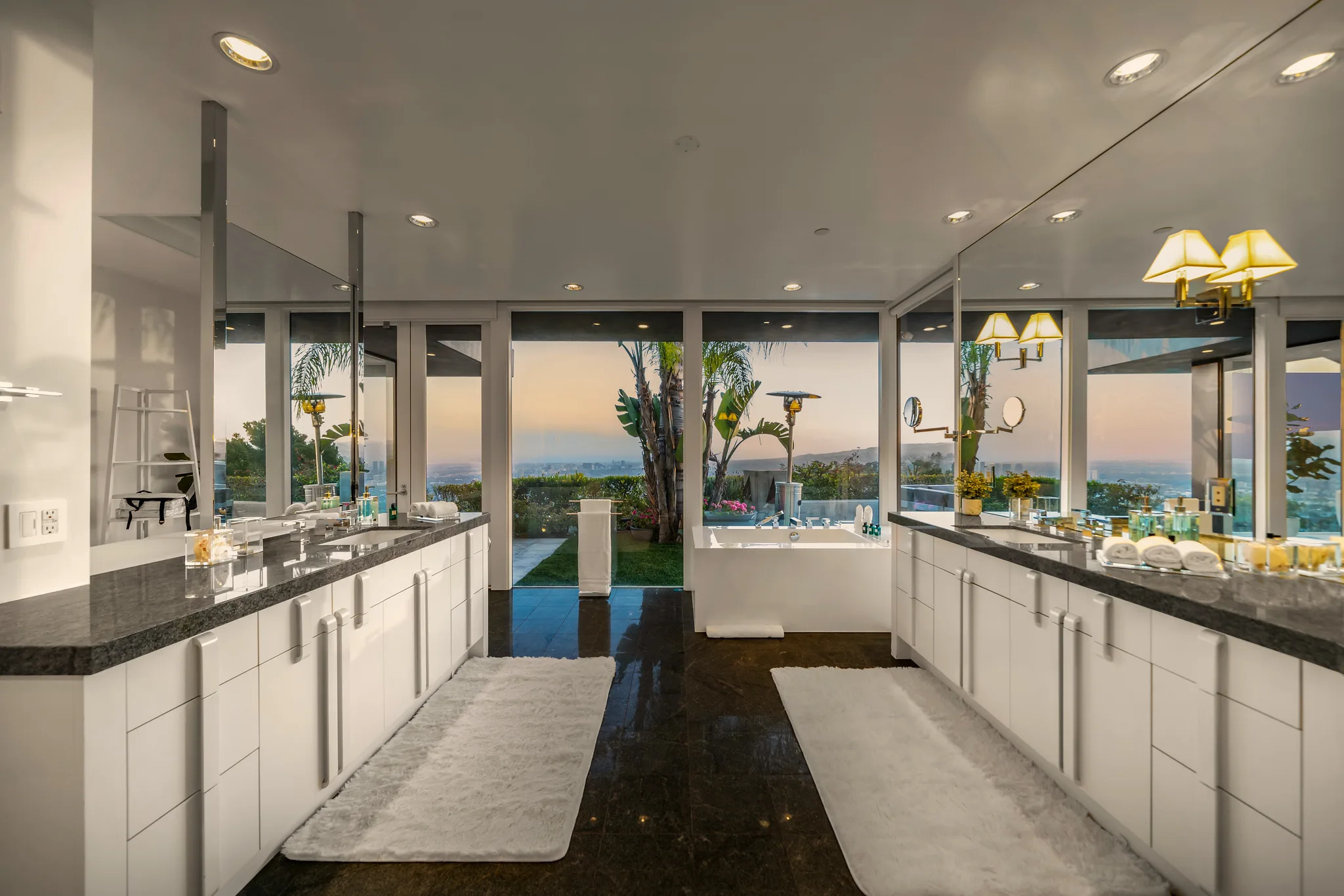 Primary bathroom with twin stone vanities, soaking tub and doors opening to a garden and distant city