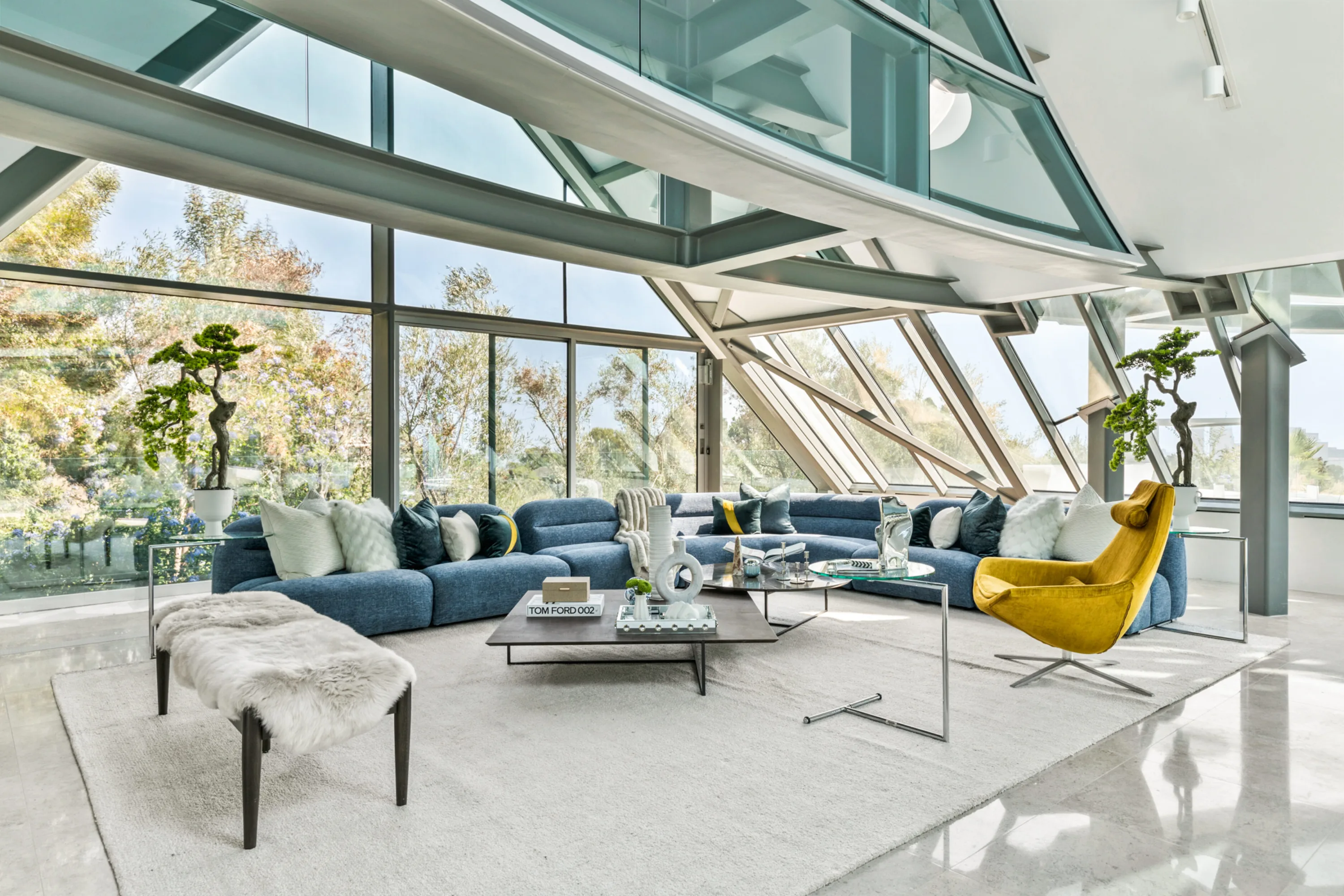 Living room under angled glass walls with blue modular sofa, mustard swivel chair and a Tom Ford 002 book