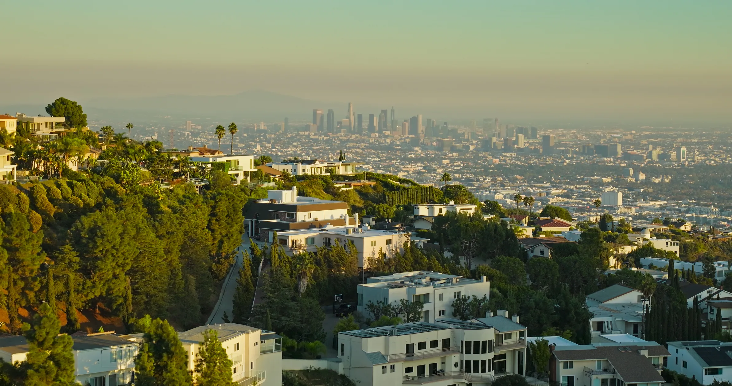 Hollywood Hills homes cascading down a ridge at golden hour with the downtown LA skyline hazy beyond