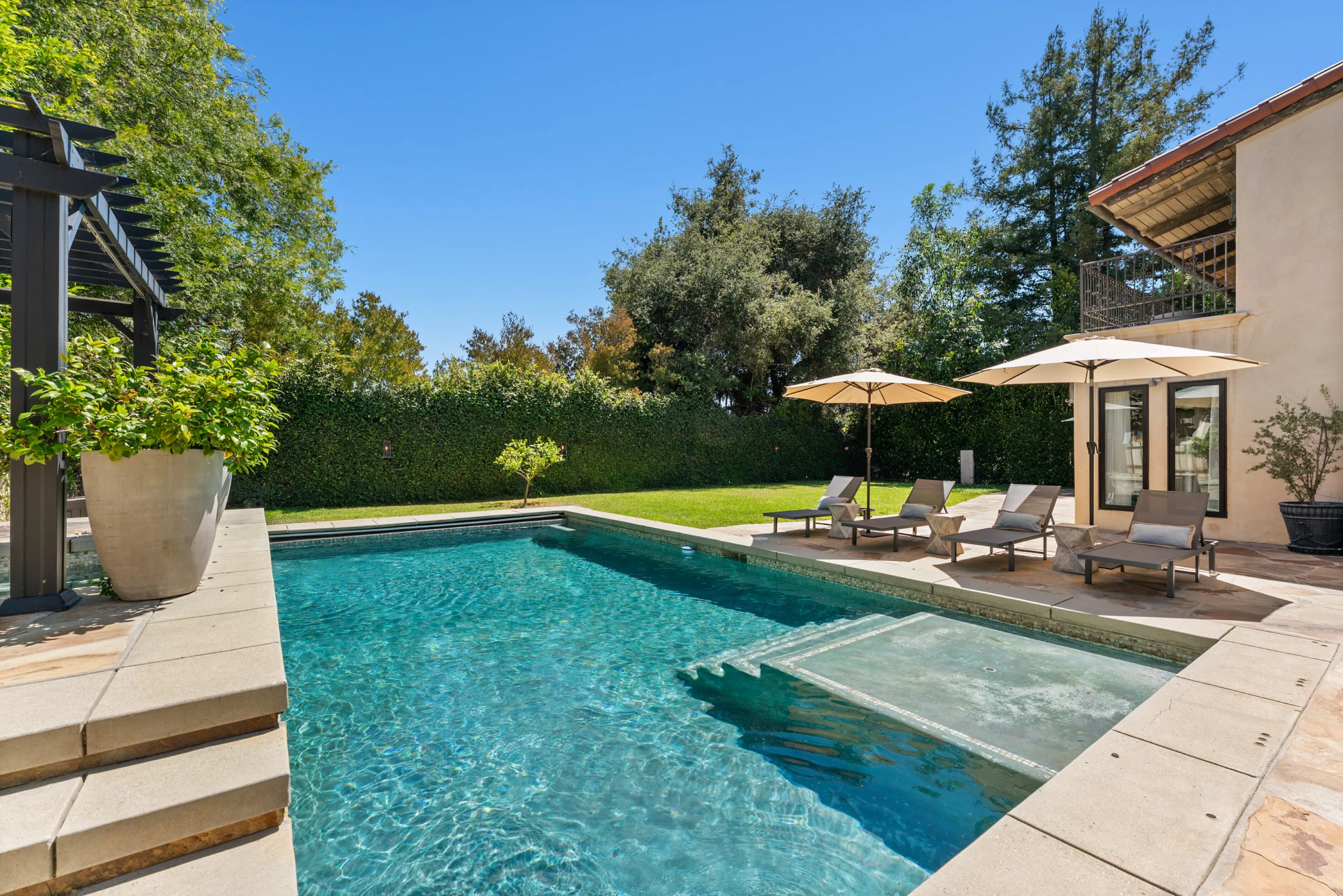 Sunlit lap pool with spa, loungers under twin umbrellas and a tall hedge for privacy