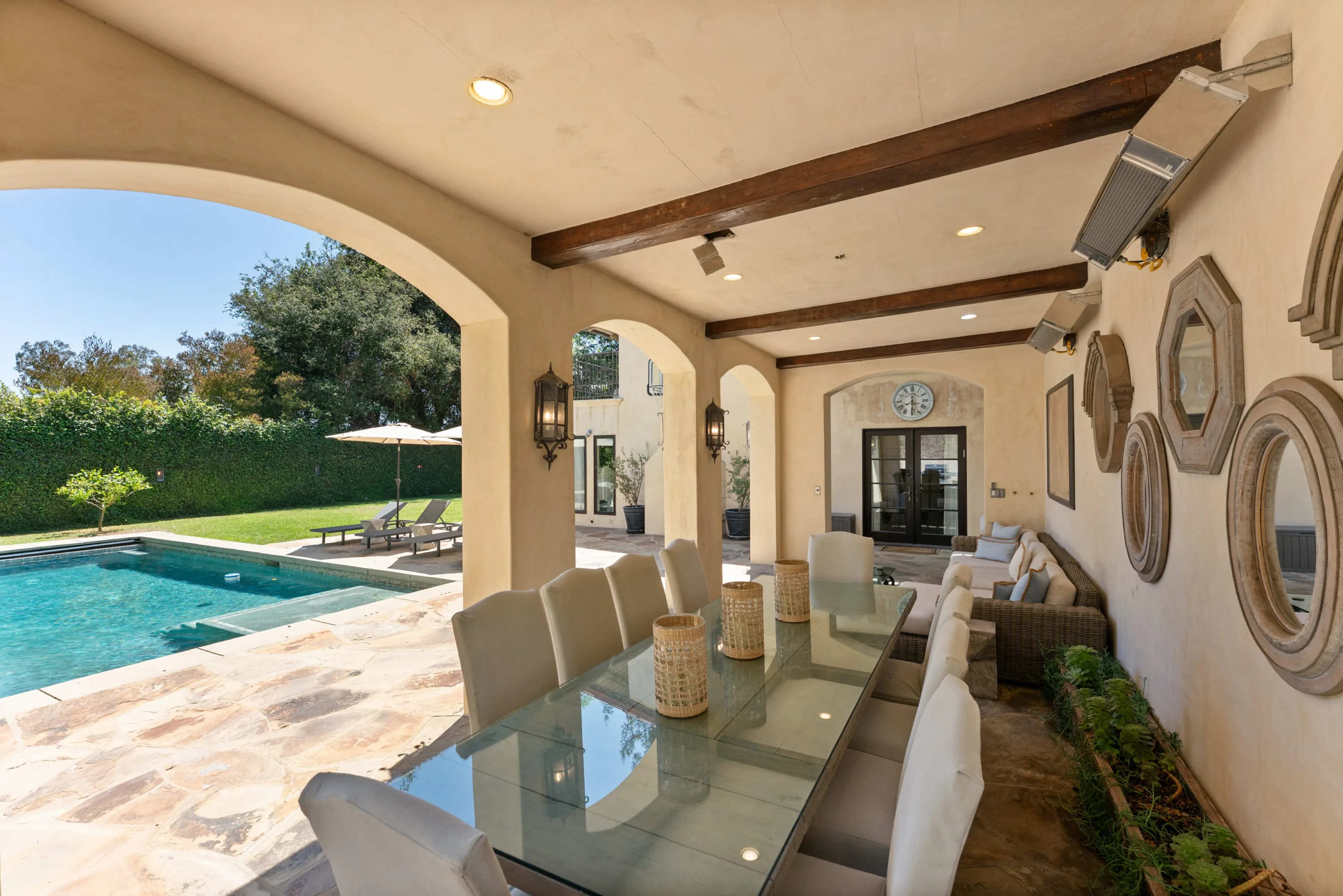 Arched poolside terrace set for ten at a glass dining table, overlooking the water and lawn