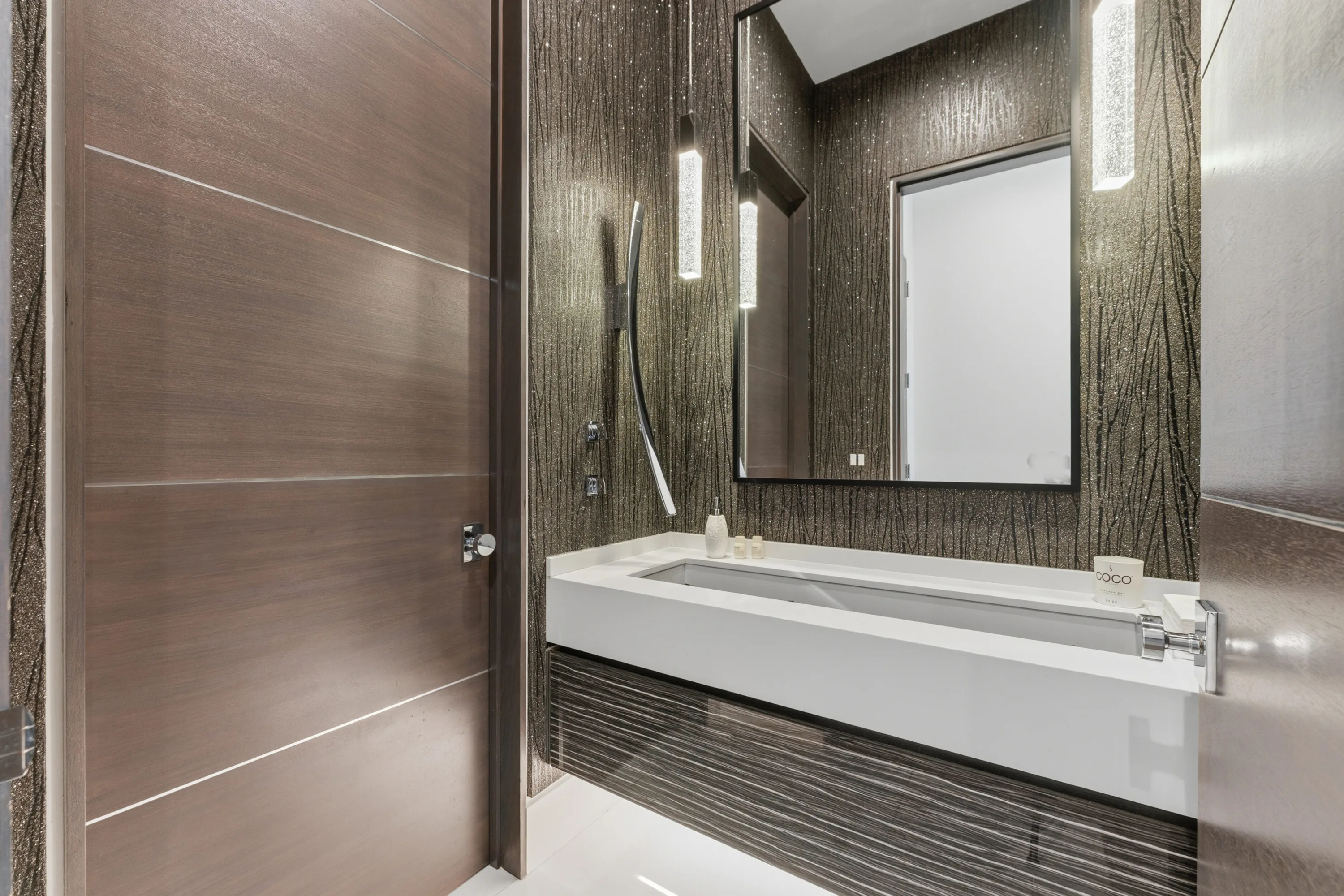 Powder room clad in textured metallic wallpaper with a long trough basin and backlit mirror