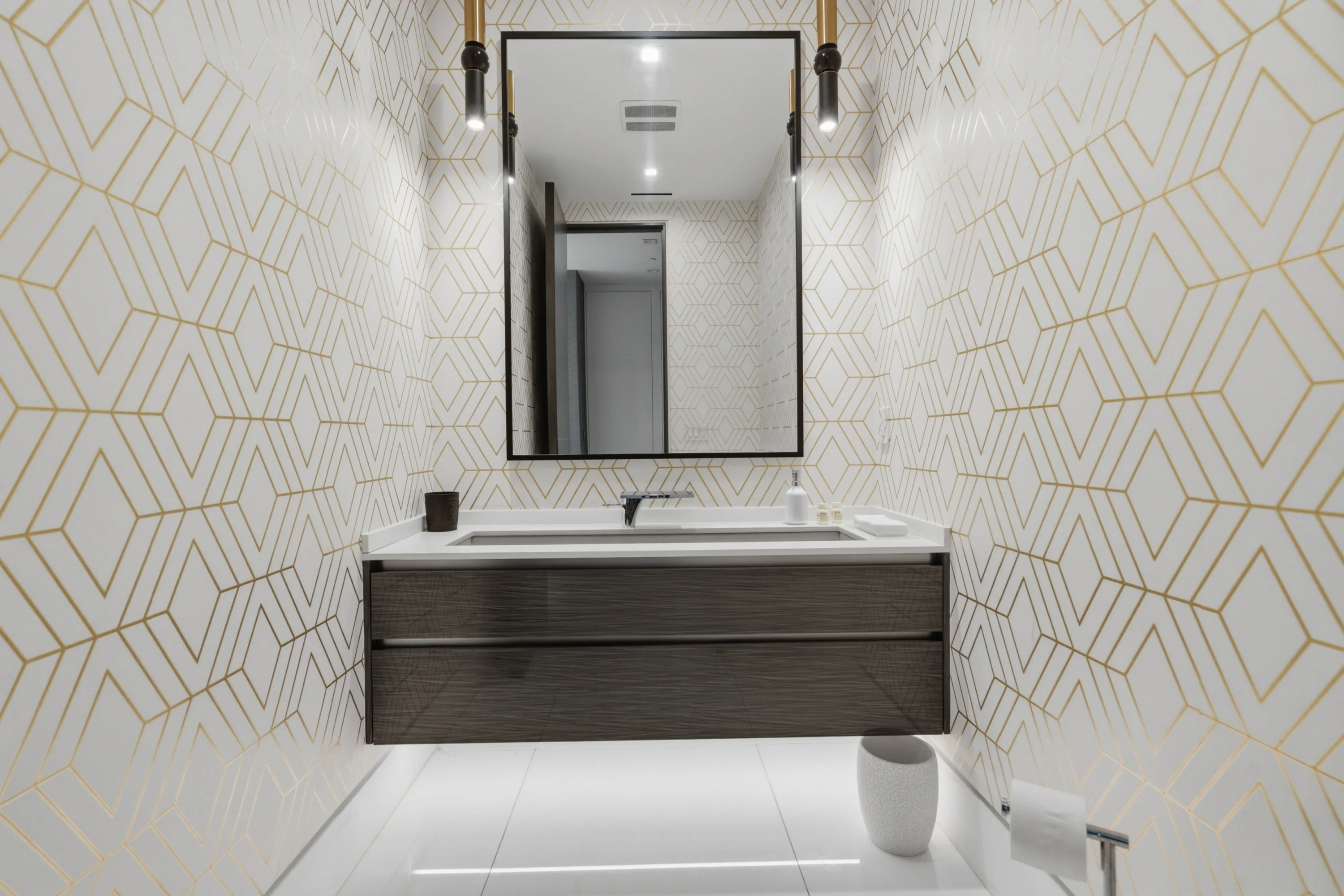 Powder room lined in white and gold geometric tile with floating dark-wood vanity and black-framed mirror