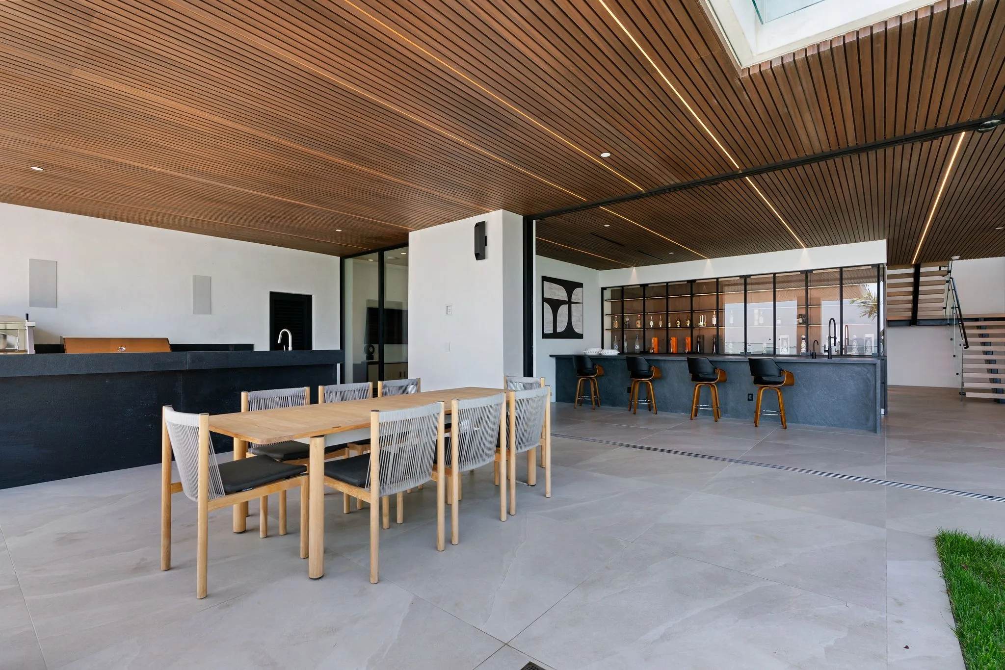 Covered terrace dining table for eight beside a black stone bar with swivel stools under a slatted timber ceiling