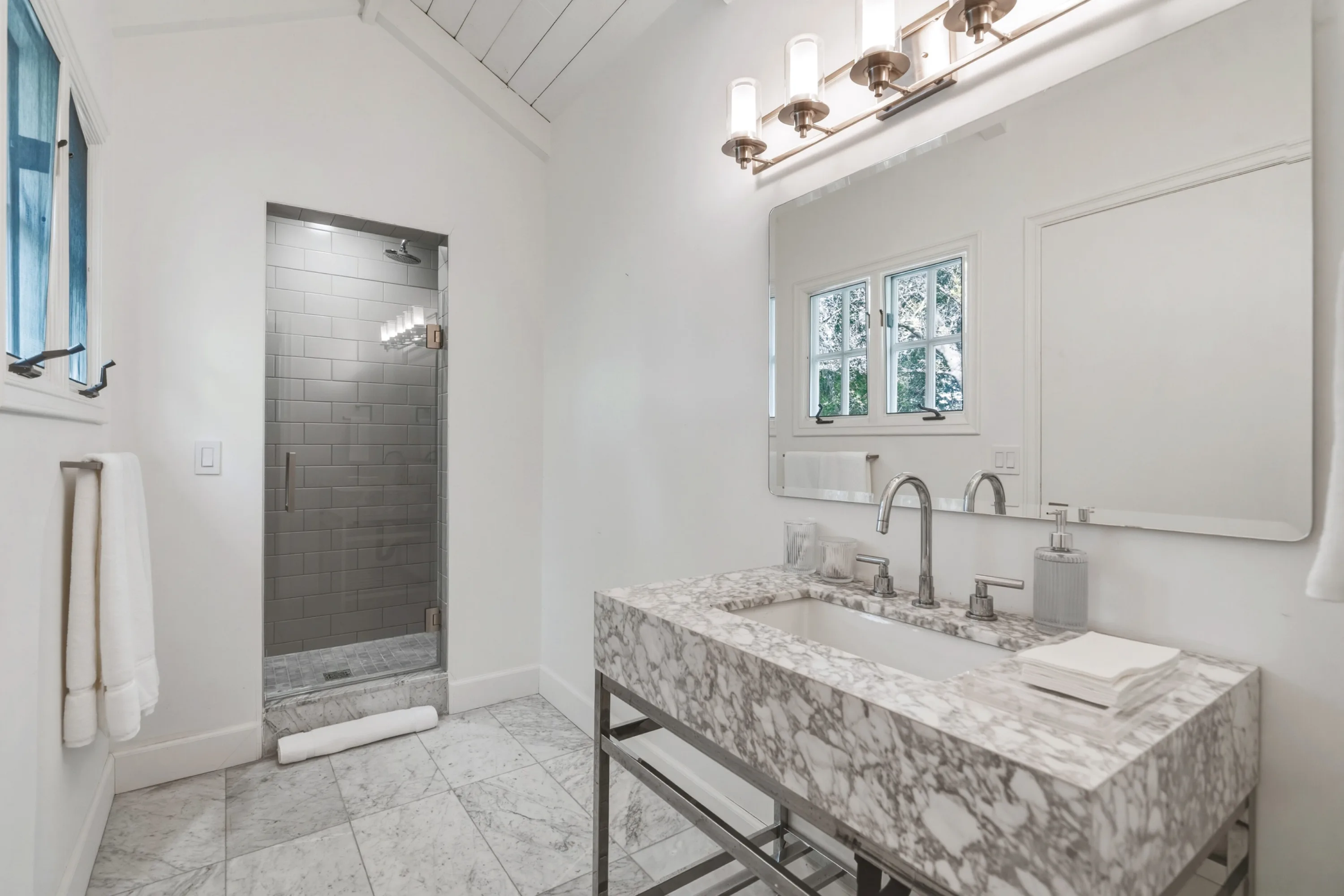 Guest bath under a vaulted plank ceiling, marble trough vanity with twin taps and a glass-enclosed grey tiled shower