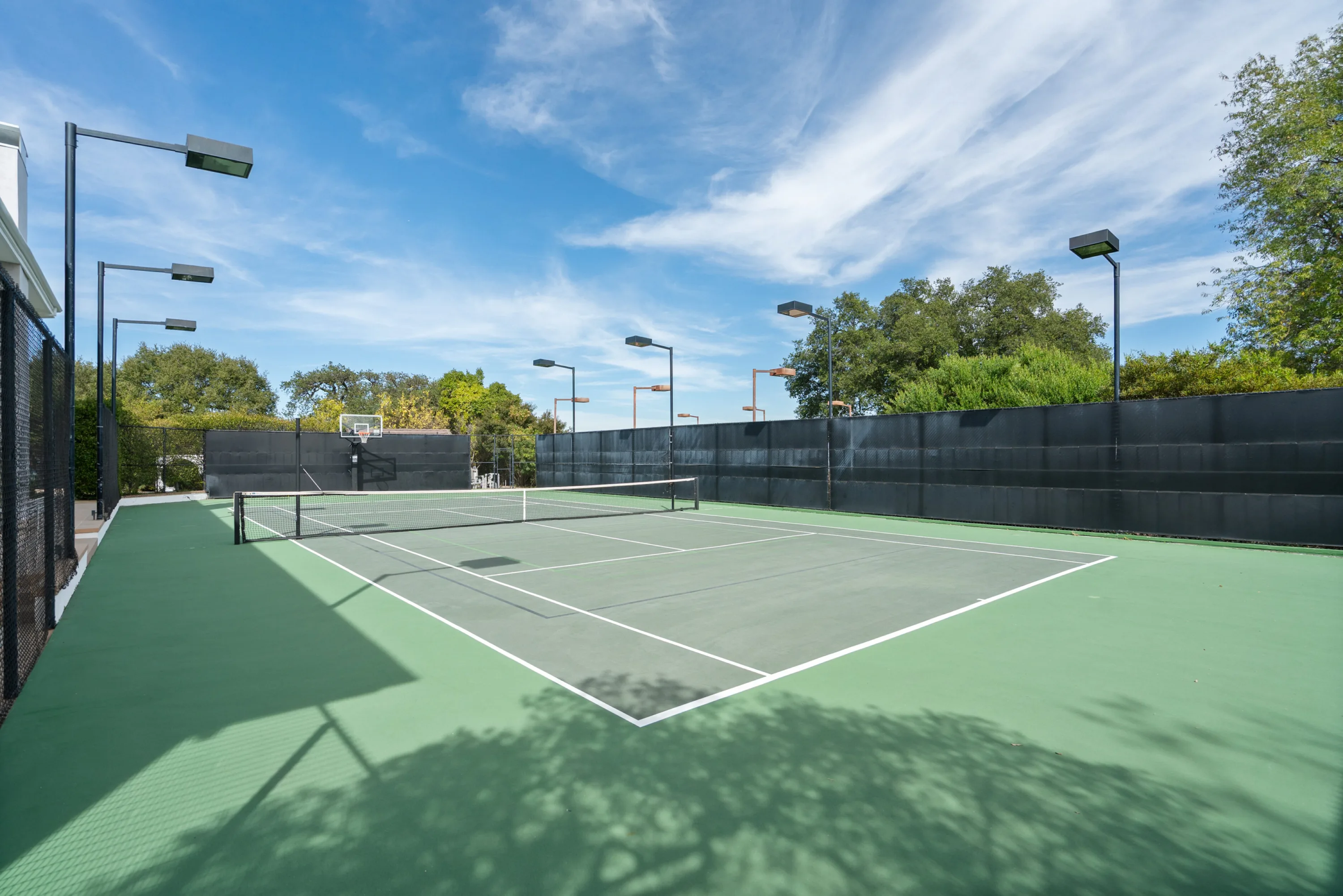 Full-size lighted tennis court with basketball hoop, hedged and ringed by floodlight poles