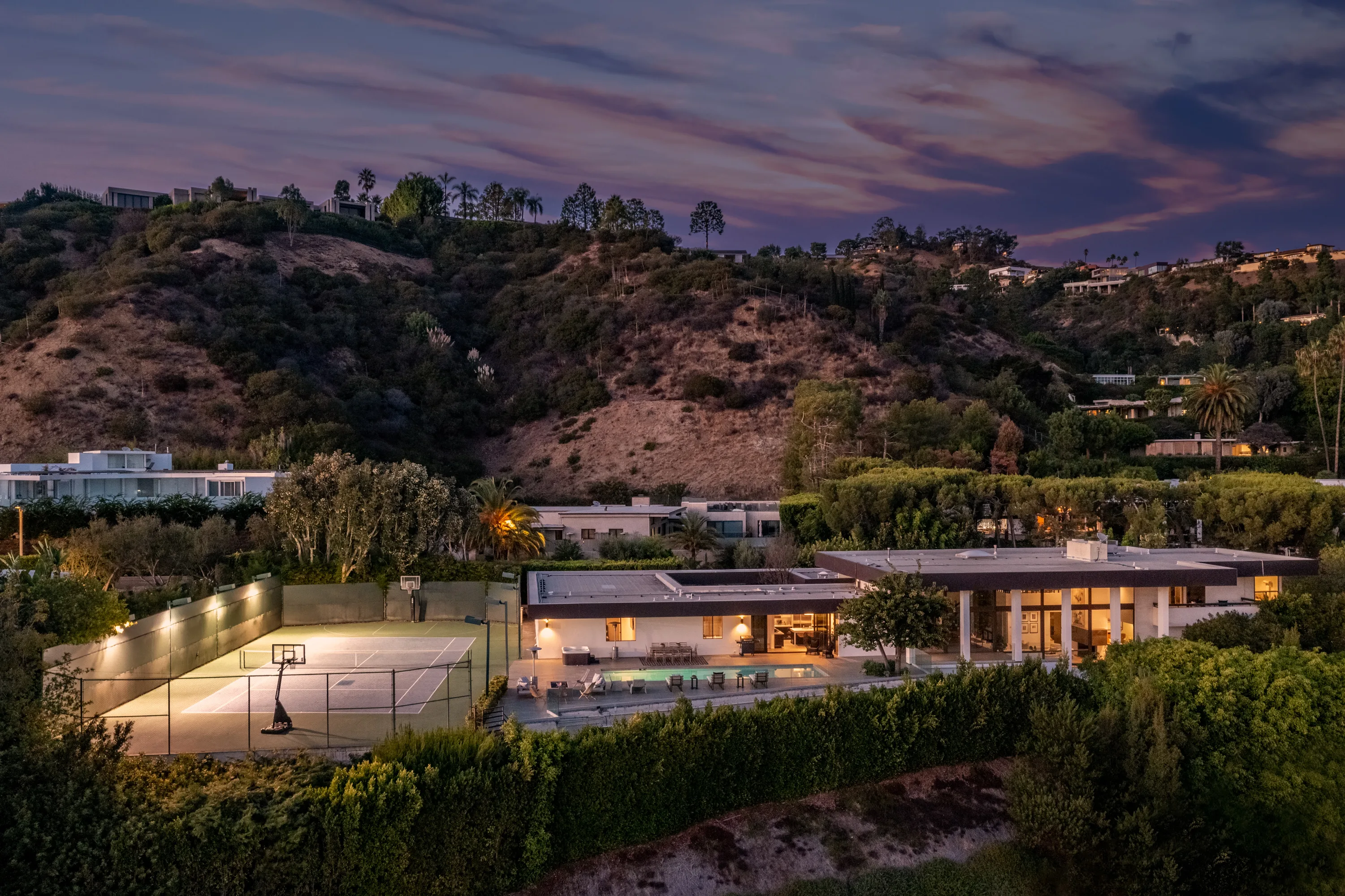 Aerial dusk view of the Trousdale Estates house with lit tennis court, pool terrace and hillside beyond