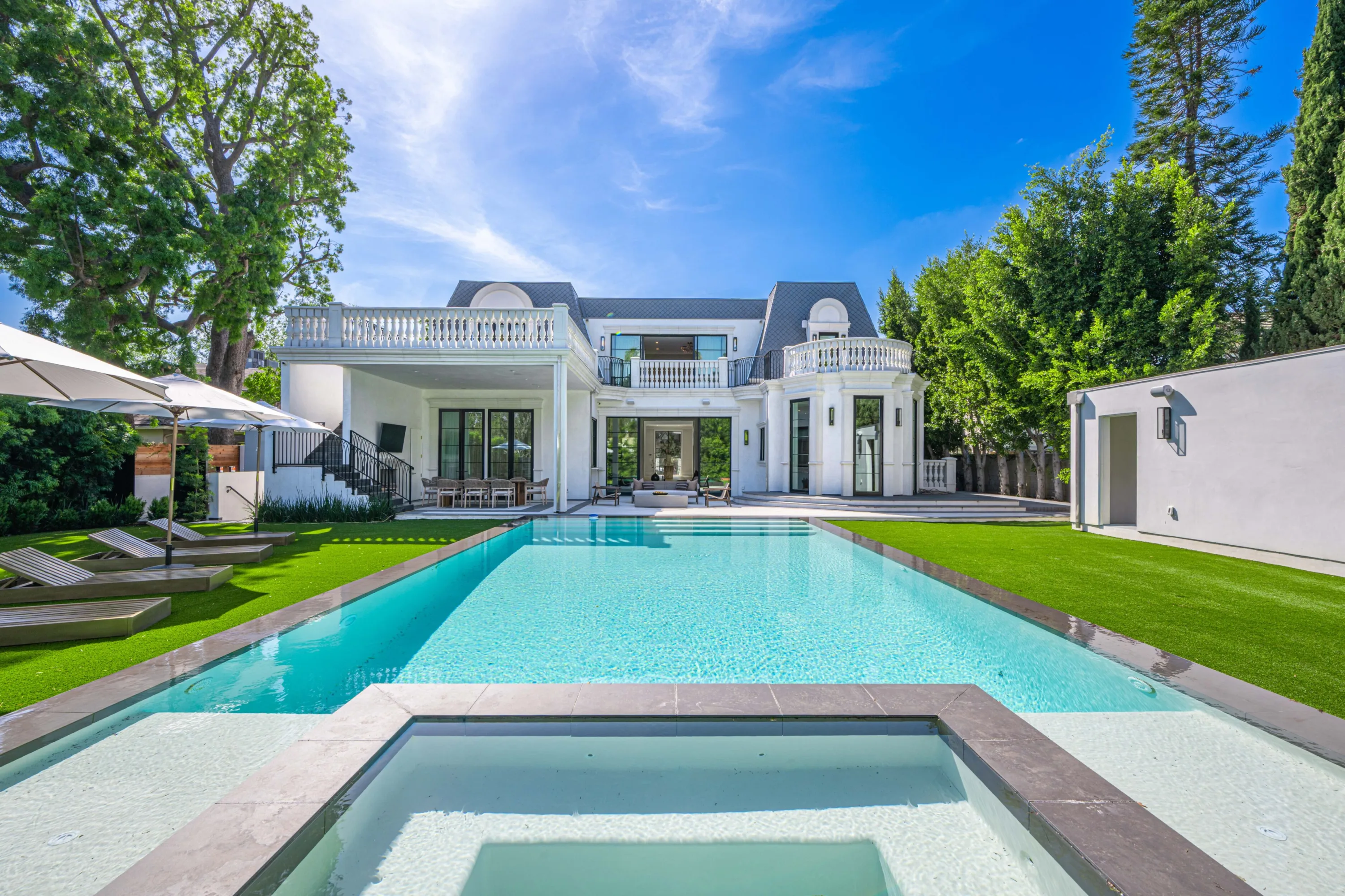 Long lap pool and sunken spa on manicured lawn, facing the estate's white rear facade and shaded loggia
