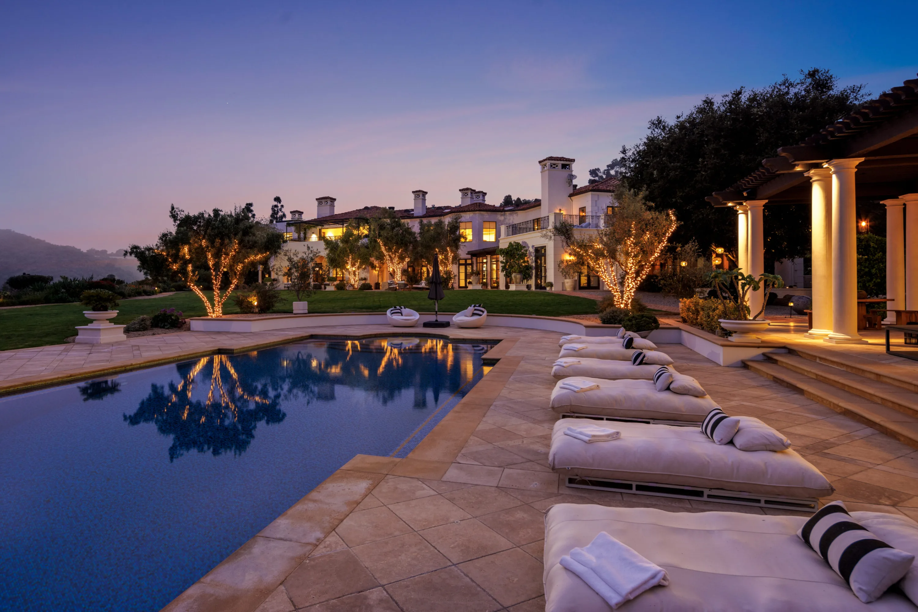 Pool and double sun loungers at dusk, with the lit Beverly Hills estate and string-lit trees mirrored in the water