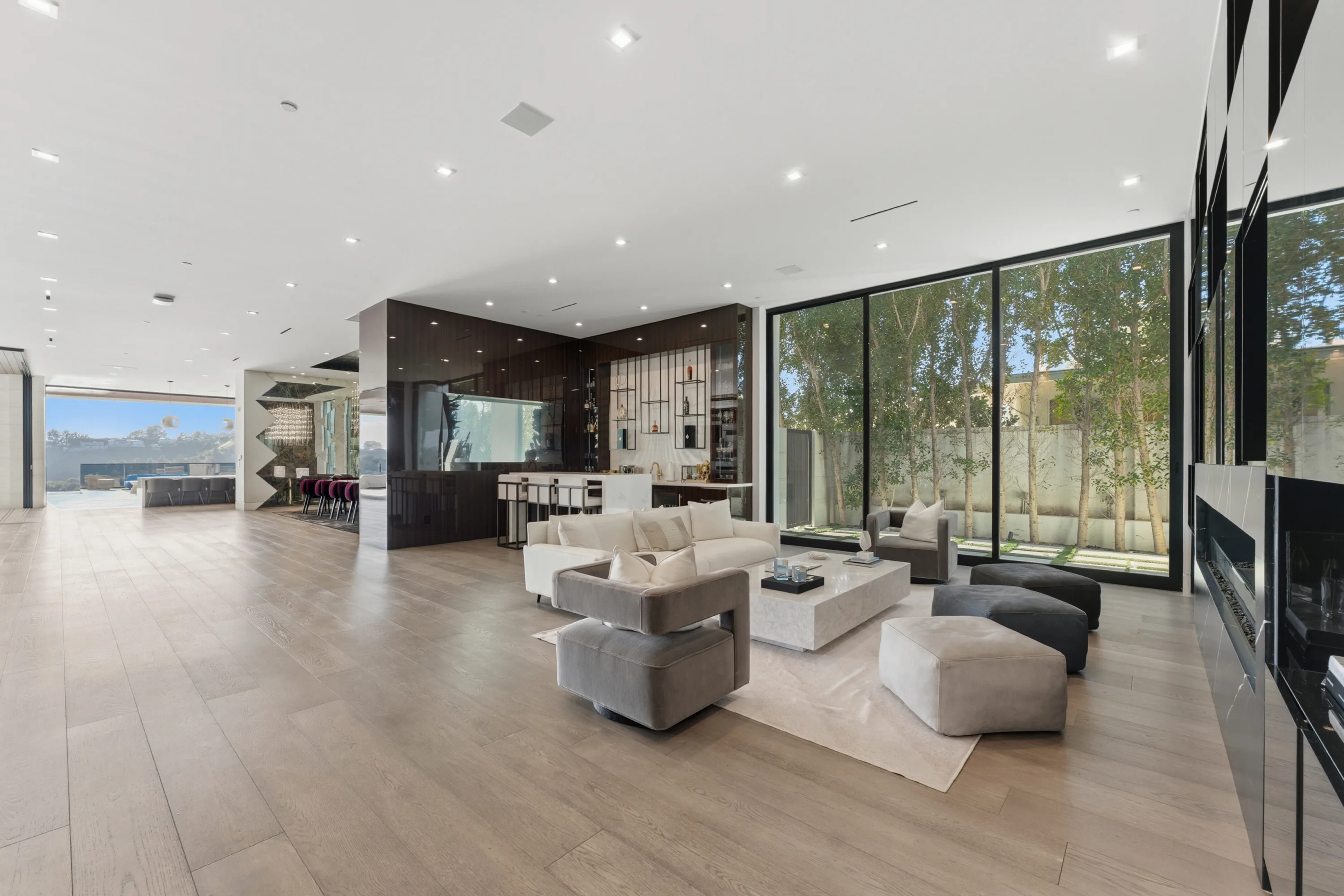 Open-plan great room in pale oak, running from linear fireplace past the bar to the pool doors beyond