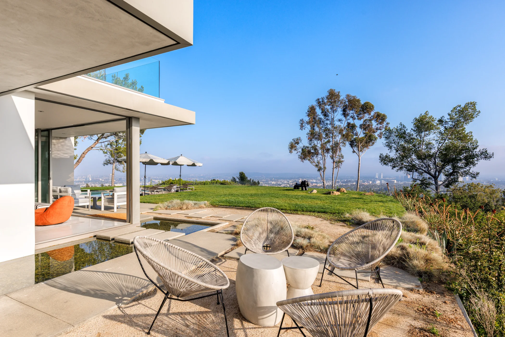 Terrace seating of woven lounge chairs beside a reflecting pool, lawn and hazy city view beyond
