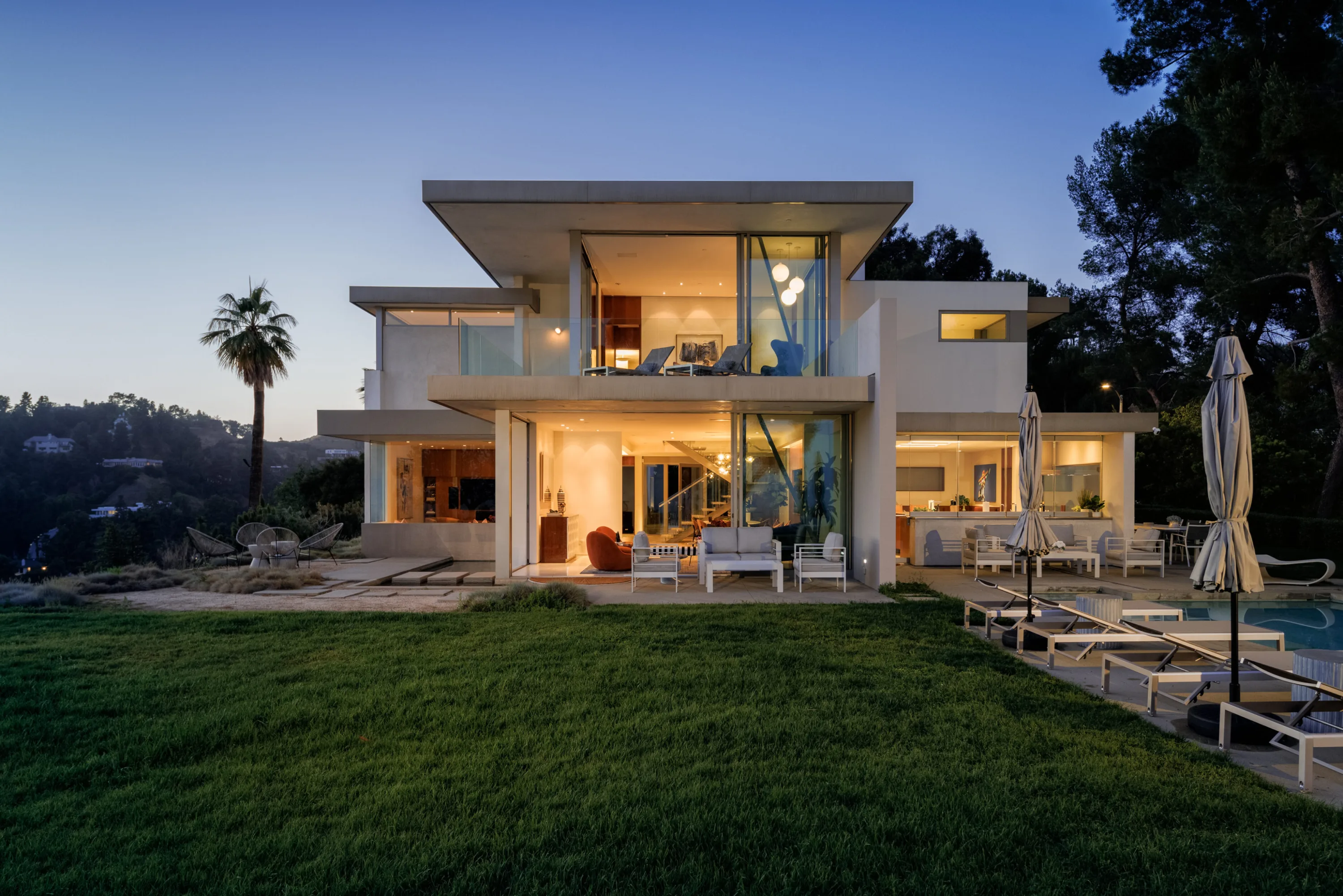 Vue Modern lit from within at dusk, glass walls open to the lawn with loungers and pool at right