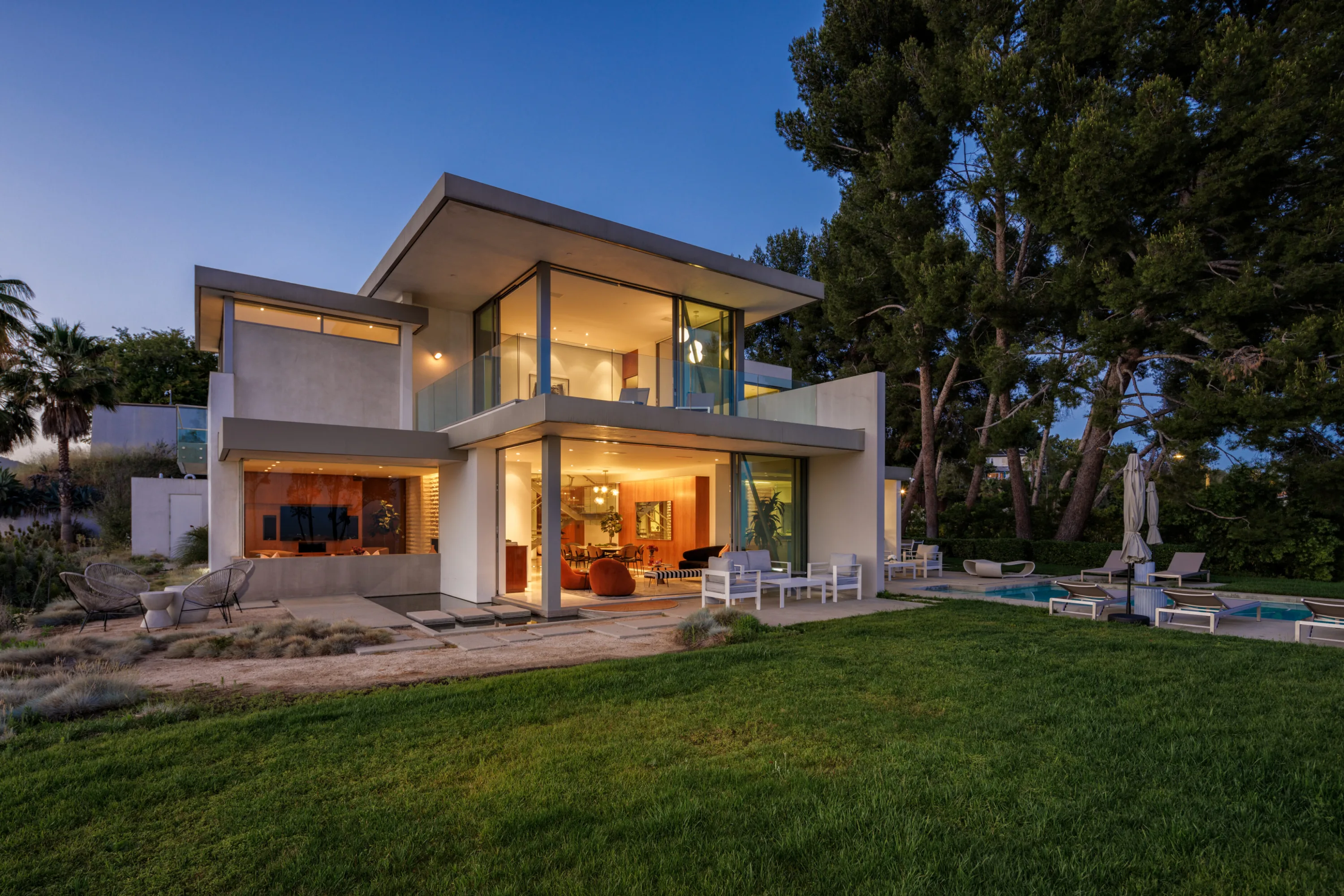 Rear elevation at blue hour, cantilevered terraces glowing above the lawn, pool and umbrellas